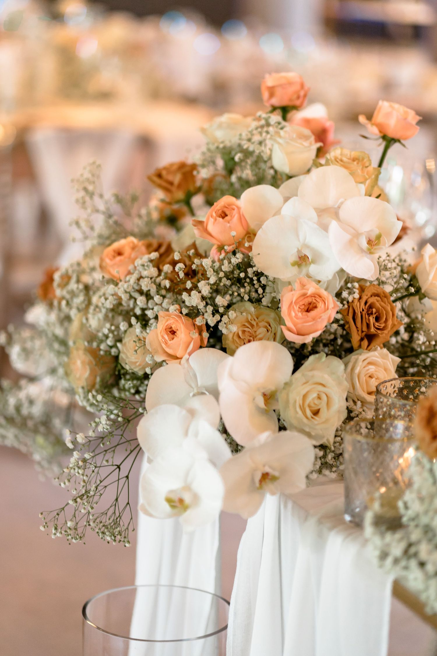 An elegant floral centerpiece with peach roses, white orchids, and baby's breath in a white vase.