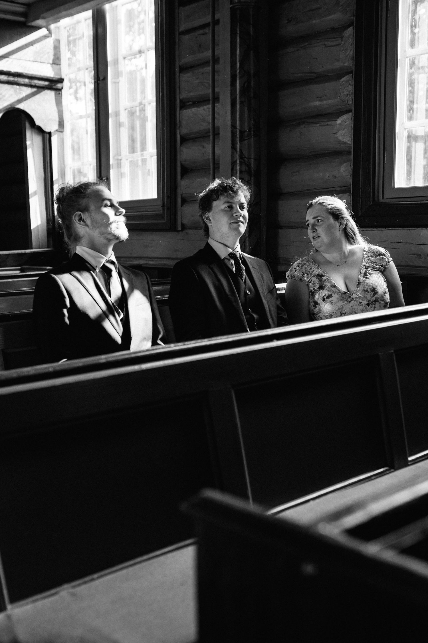 Wedding guests seated in wooden church pews illuminated by dramatic side lighting.
