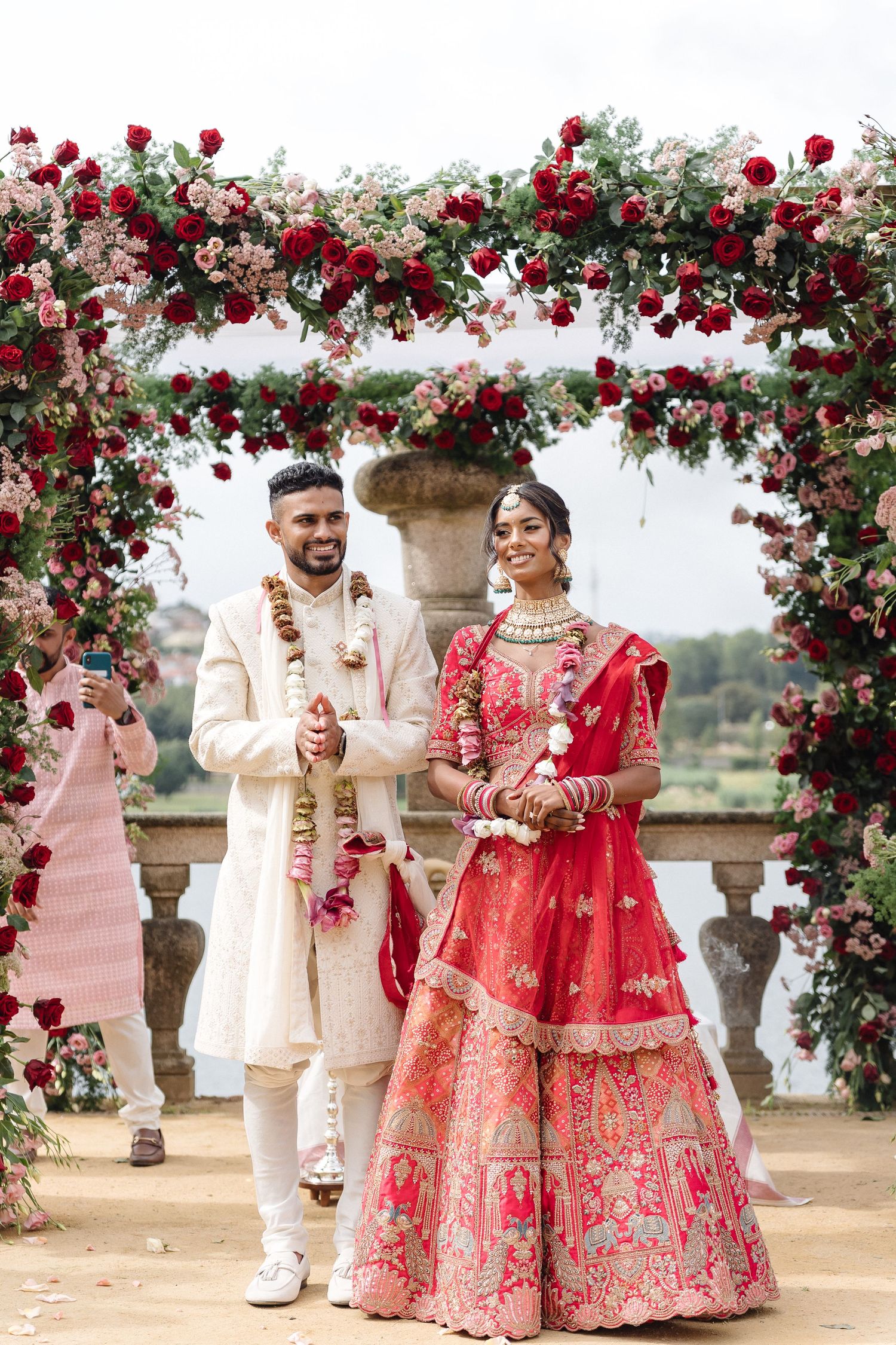 Elegant wedding arch decorated with red roses and white pillars in outdoor setting.