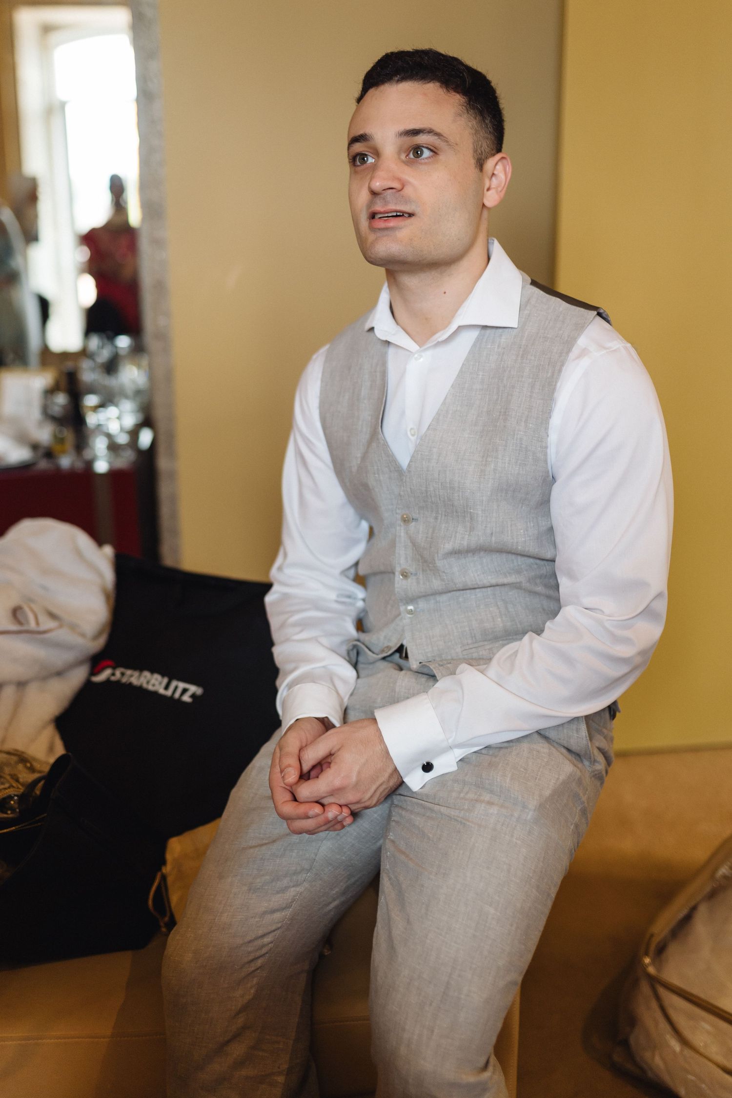 Groom in formal grey vest and white shirt sitting on couch during wedding preparations.