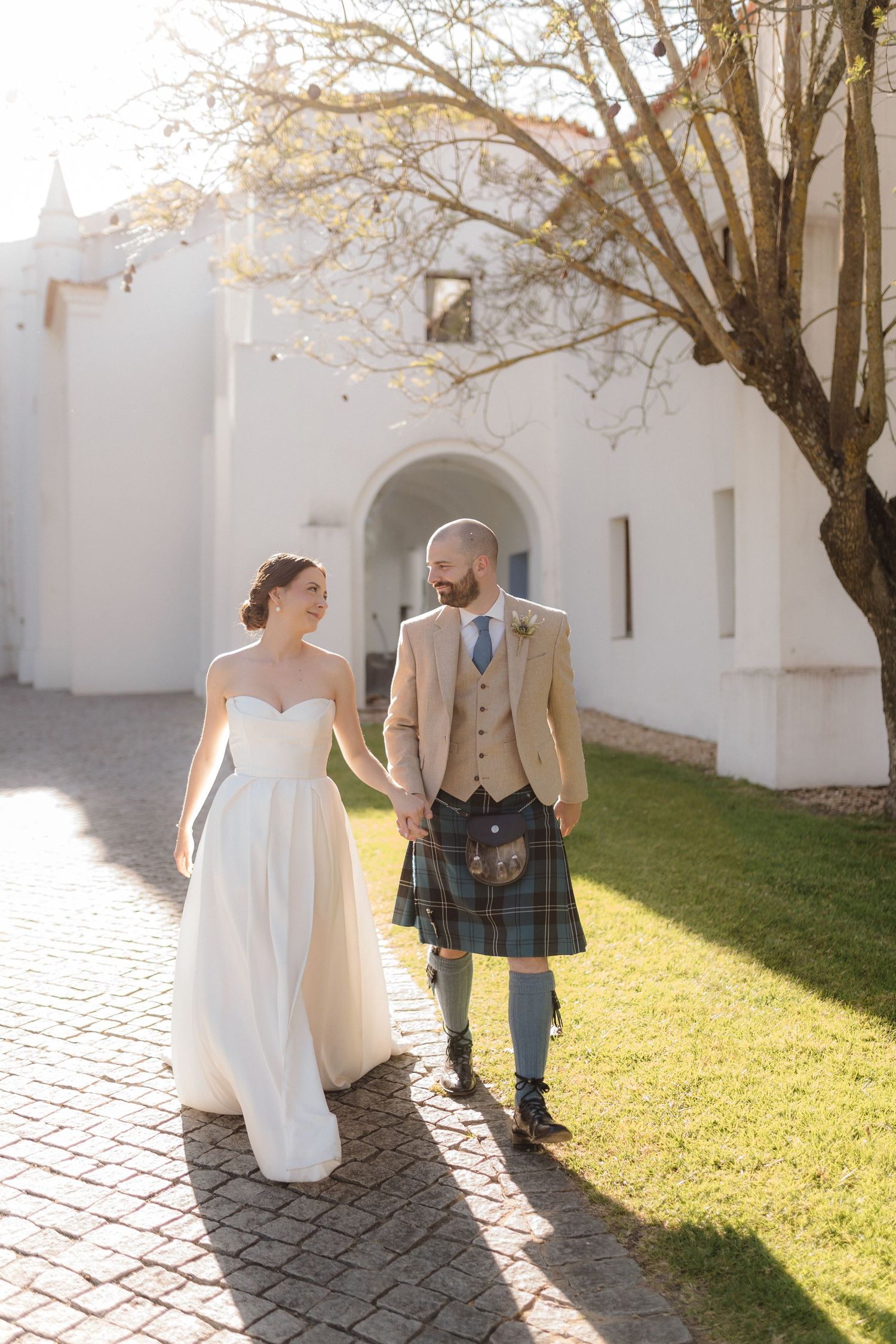 A couple in wedding attire walks along a white stone path on a sunny autumn day with trees casting shadows.