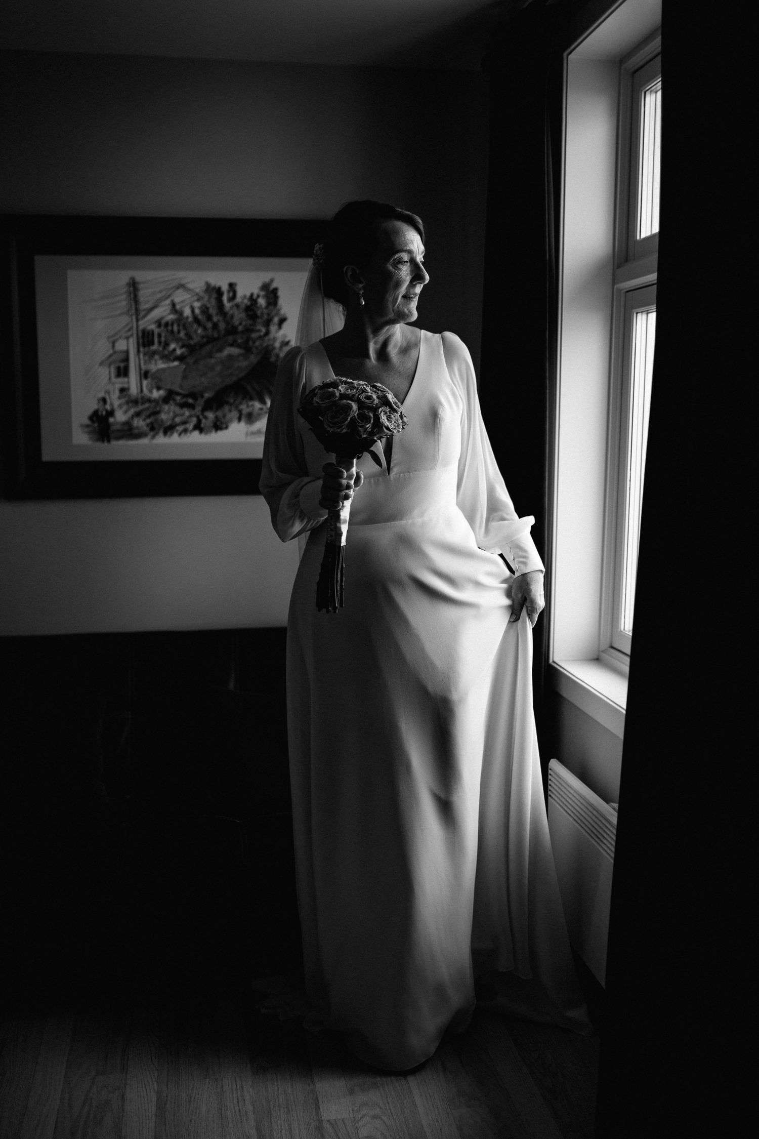Black and white photos of a bride in a flowing white dress standing by a window in dramatic lighting.