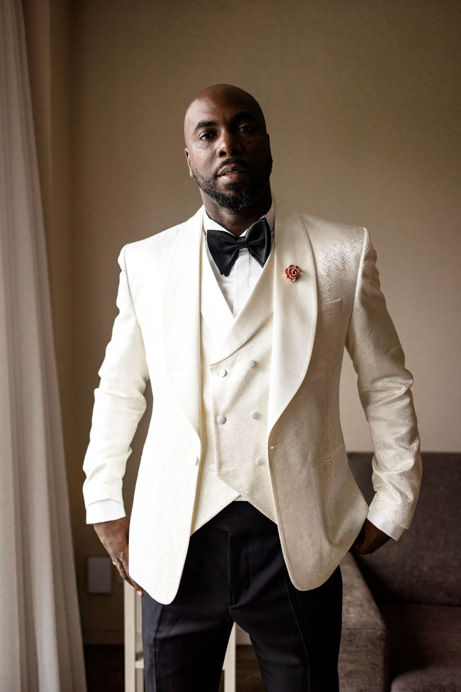 A formal portrait of a groom wearing a white tuxedo jacket with black bow tie and pants.