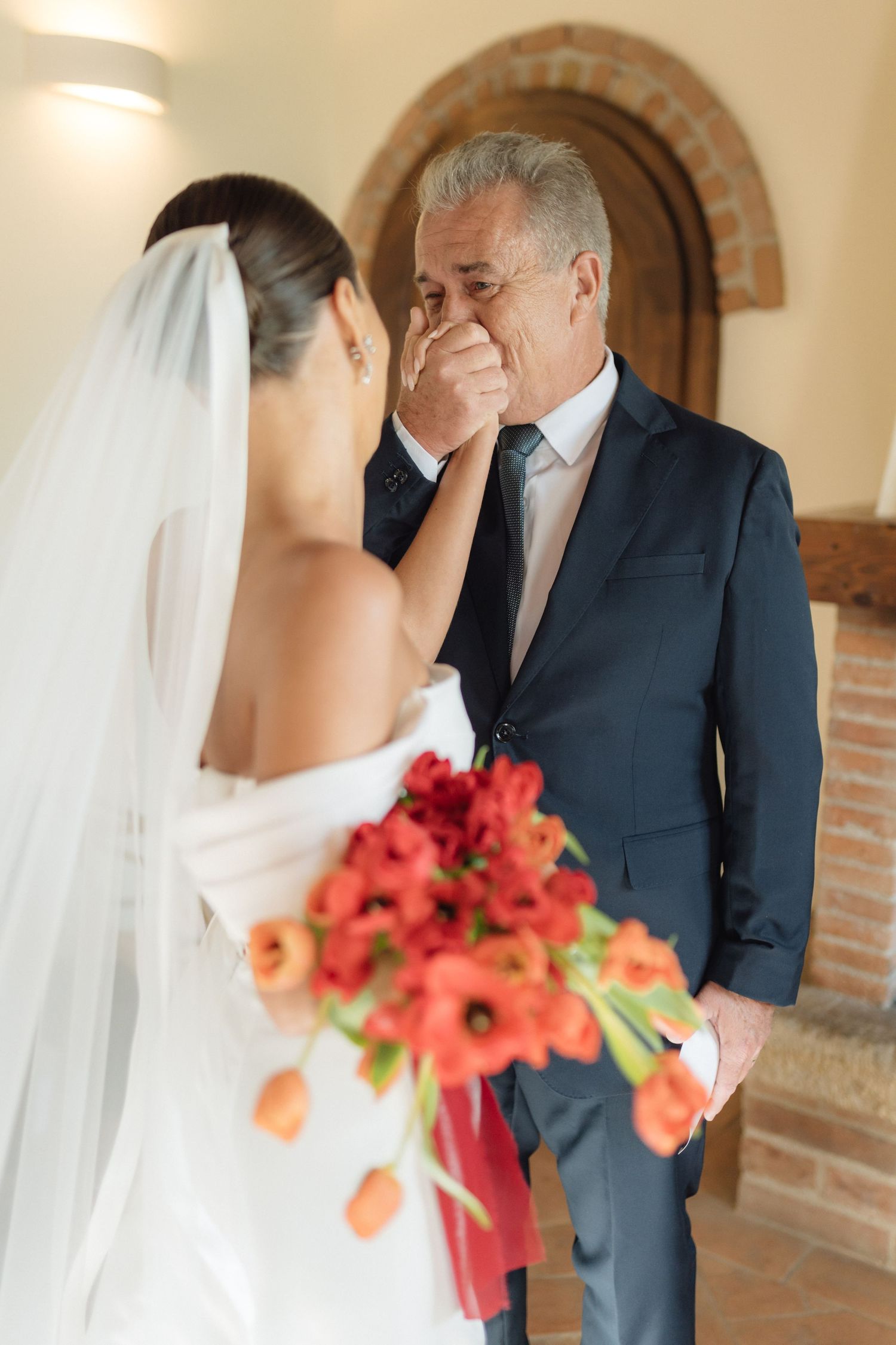 Emotional father wiping tears seeing bride in white dress holding red tulip bouquet during wedding first look.