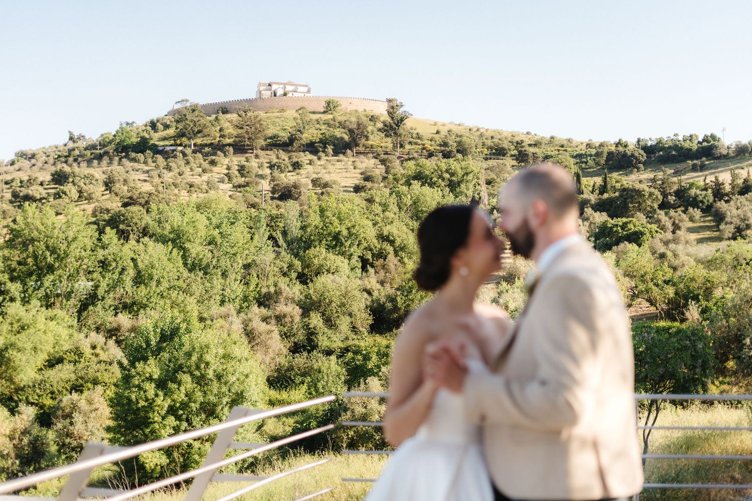 A wedding couple embraces in a sunlit outdoor setting with mountains in the background.
