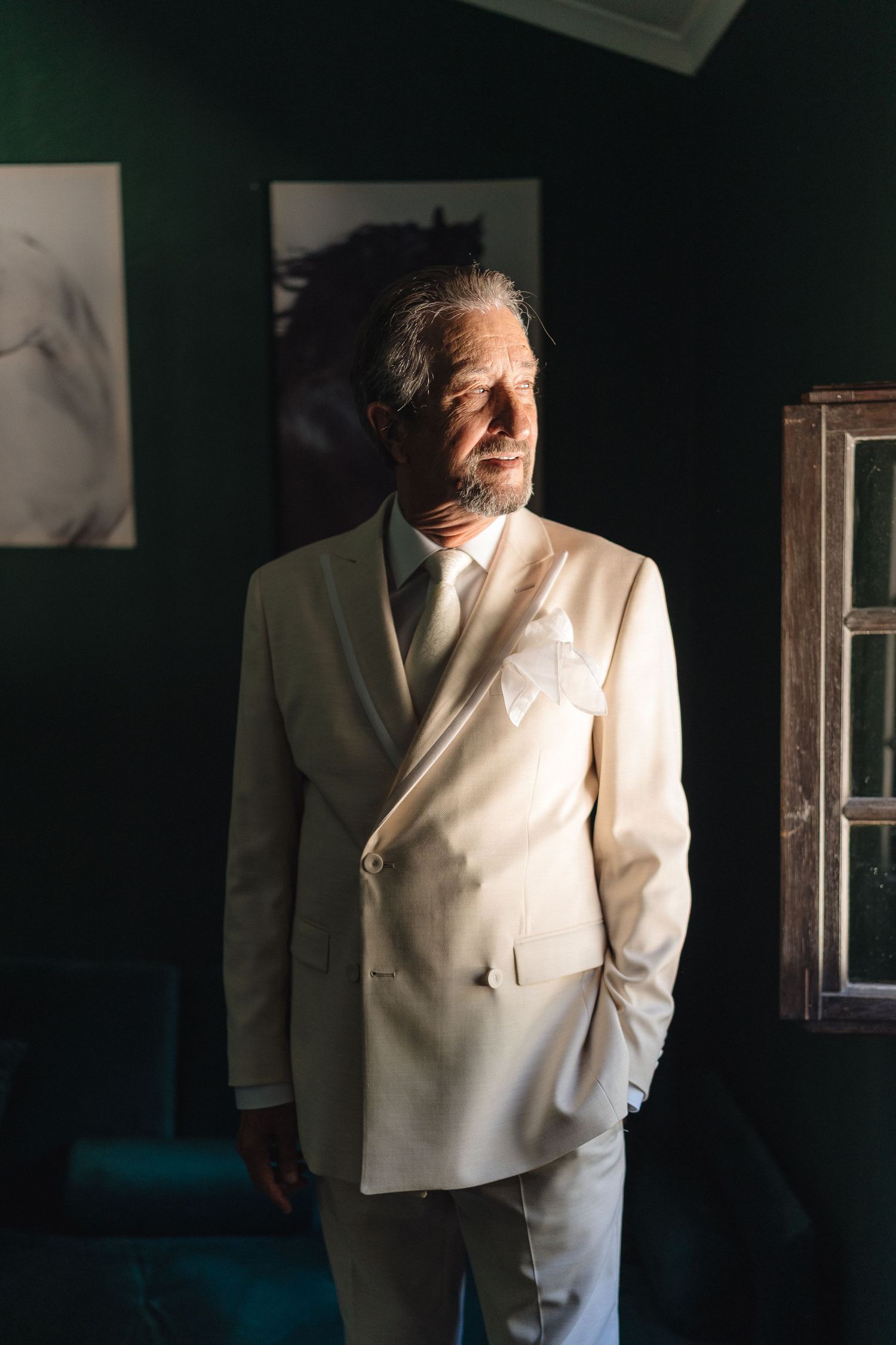 Person in a cream colored suit stands in a dimly lit room with dramatic shadows.