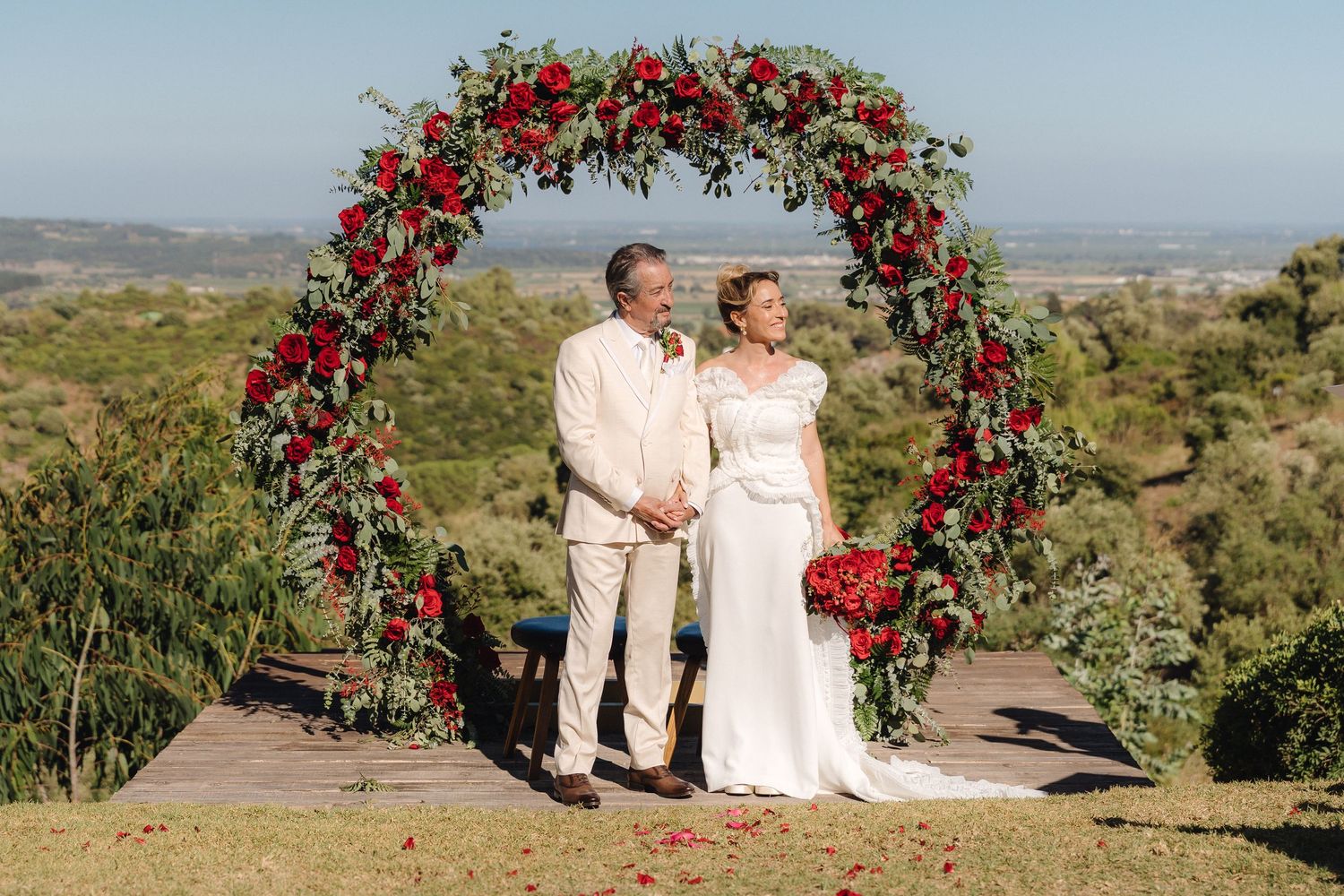 Beautiful floral arch with red roses frames a scenic outdoor wedding ceremony.