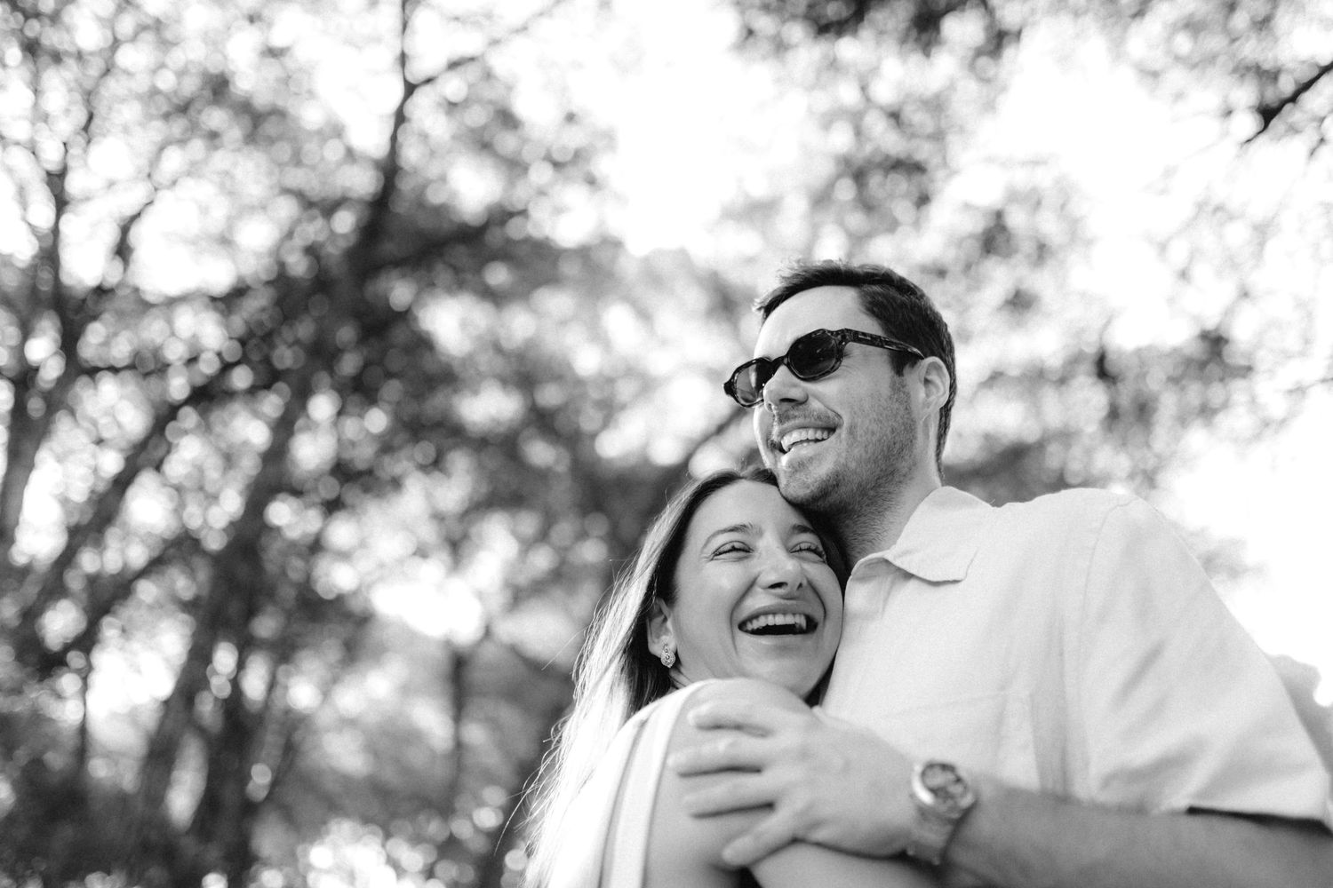 Happy couple laughing and embracing outdoors in black and white photo with trees in background.