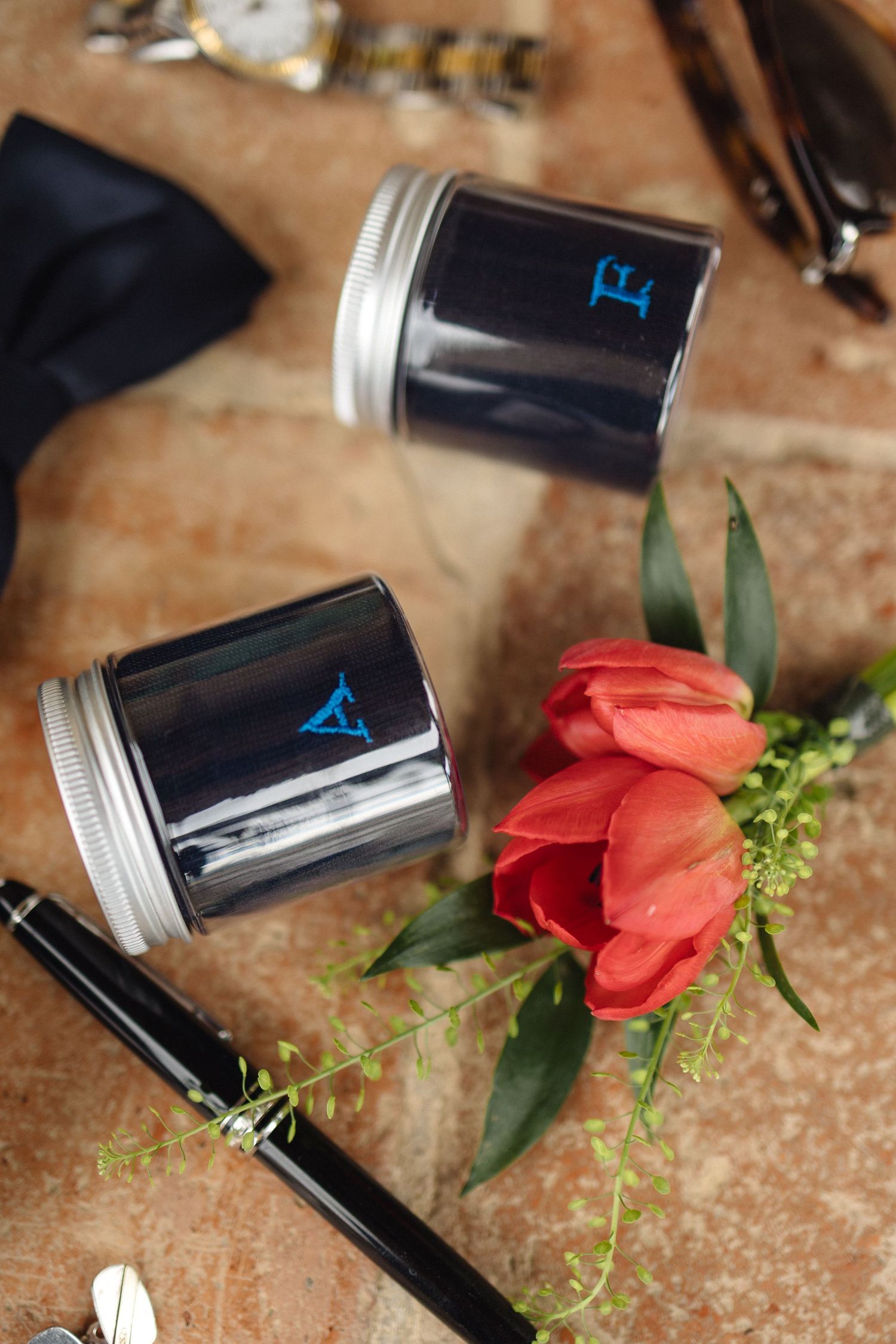Two dark ink jars with blue monograms beside red tulips and a pen on a stone surface for a wedding.