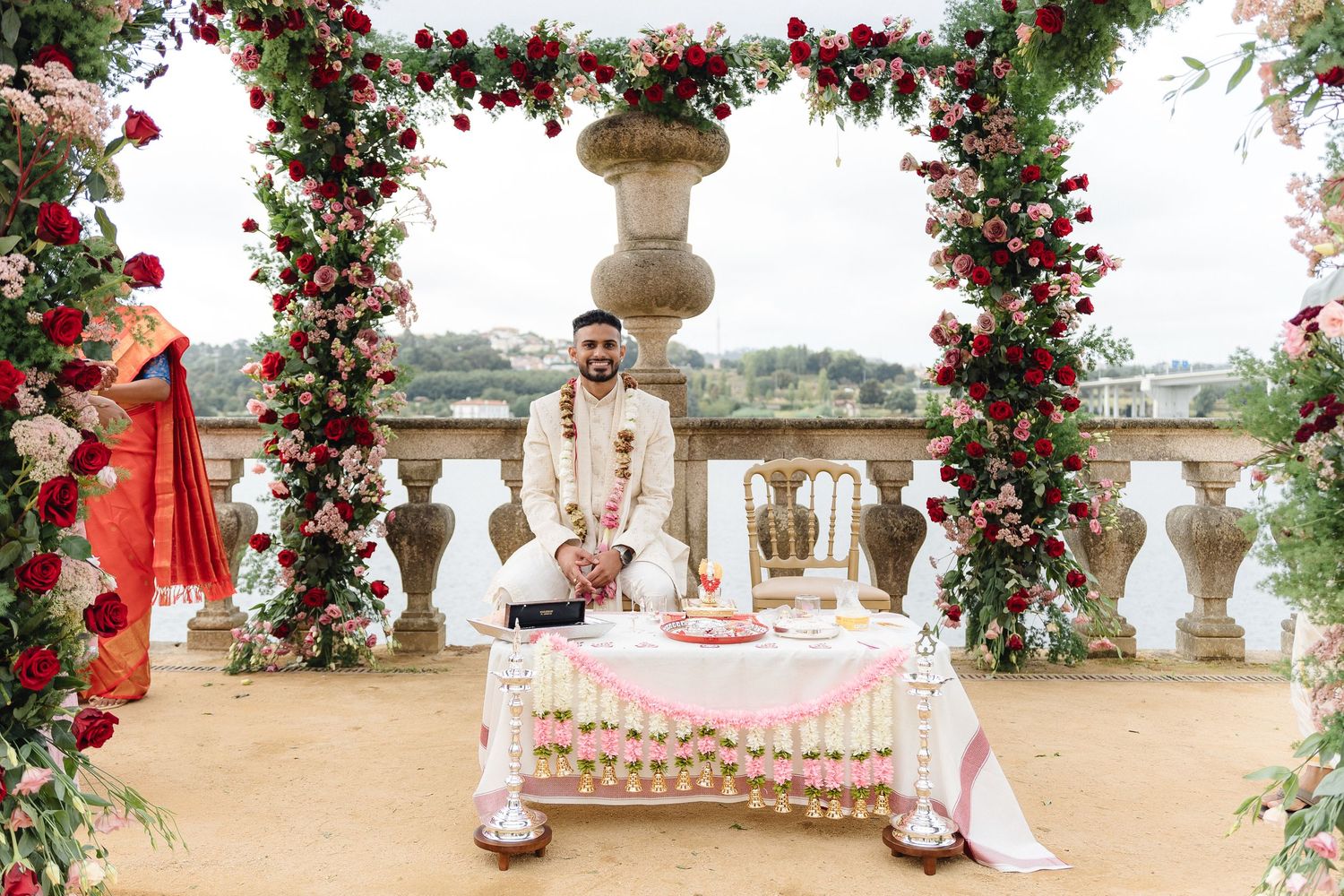 Wedding ceremony setup with red floral arches and white chairs along a waterfront balcony.