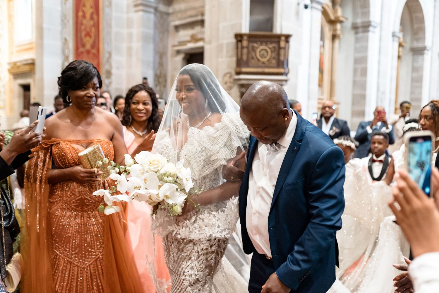 Wedding party walks down church aisle with bride in lace gown holding white bouquet.
