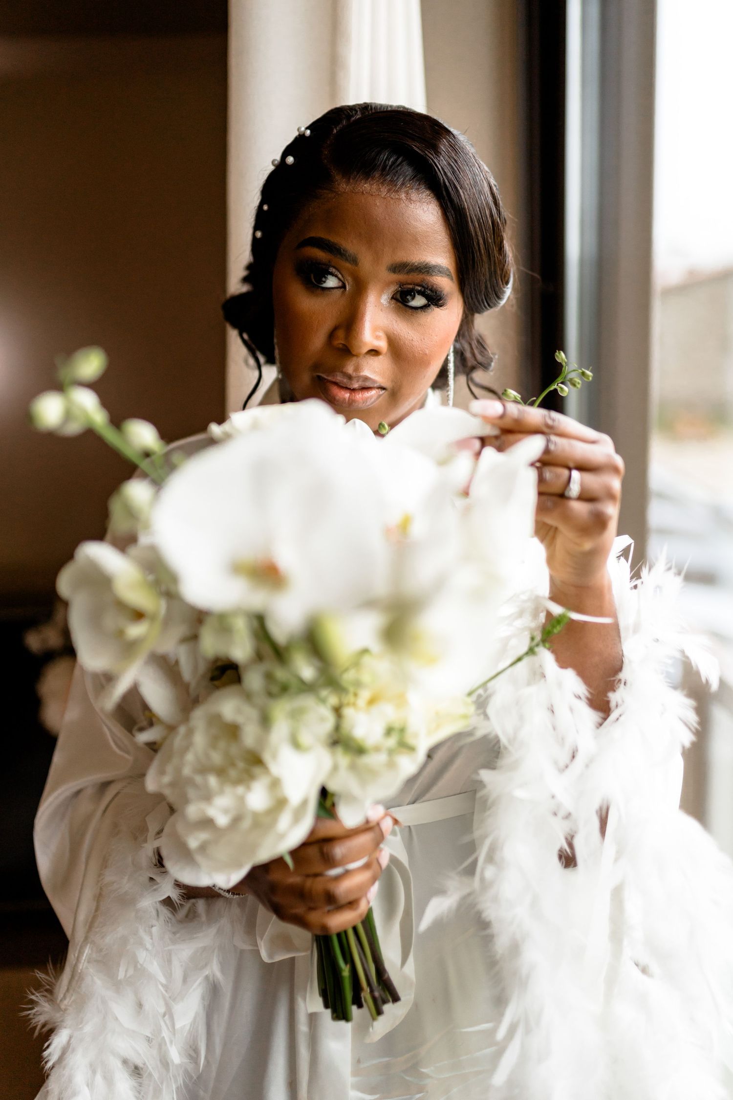 A bride in white holds an elegant bouquet of white flowers and greenery near a window.