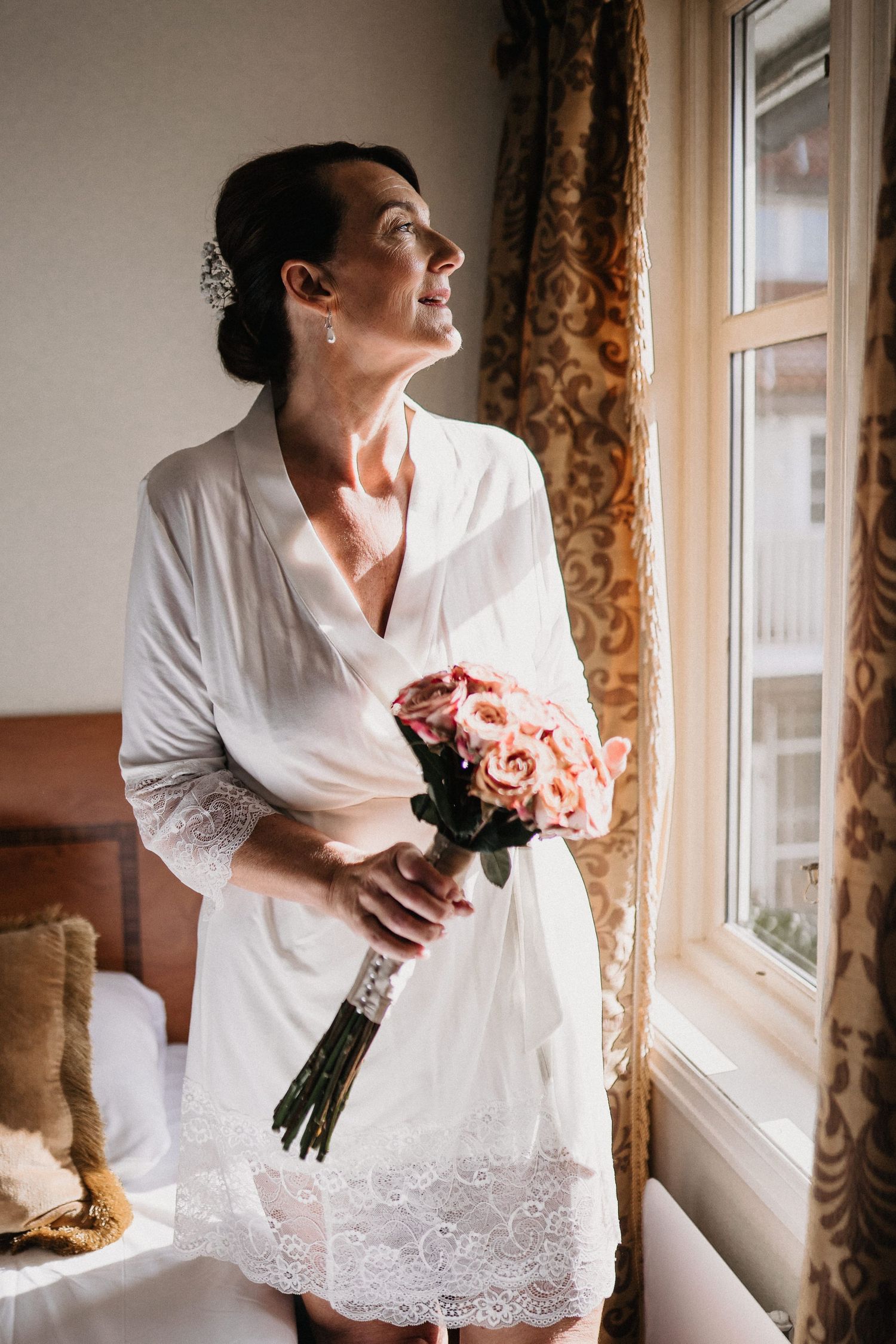 A person in a white lace robe holds a bouquet of pink roses while standing near a window with ornate curtains.