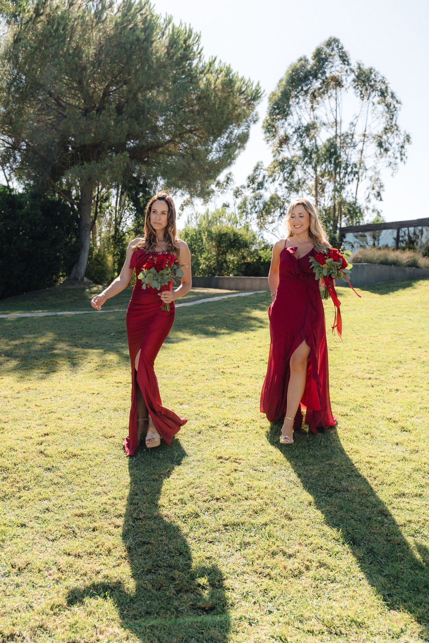 Two figures in elegant burgundy dresses with thigh slits pose on a grassy lawn with trees in the background.