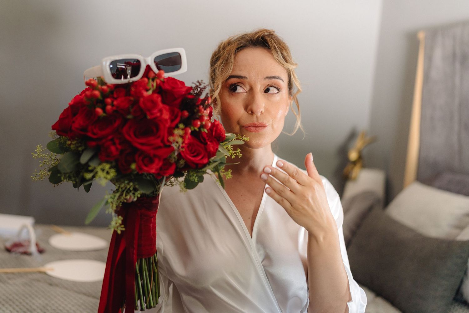 Person in white wearing sunglasses with red rose bouquet on bed.