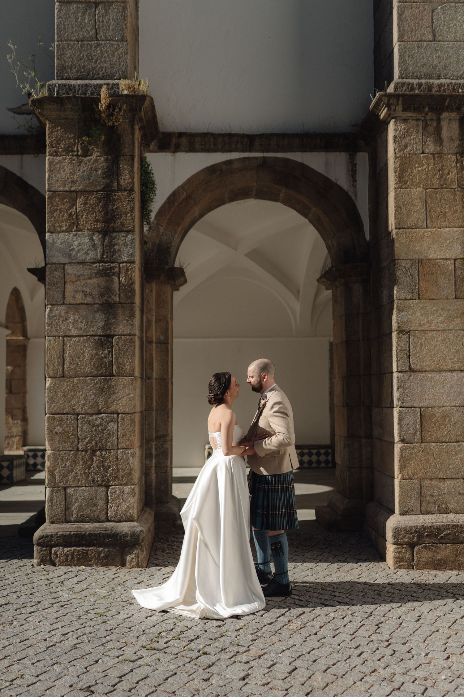 A wedding couple poses under stone archways in a historic building courtyard.
