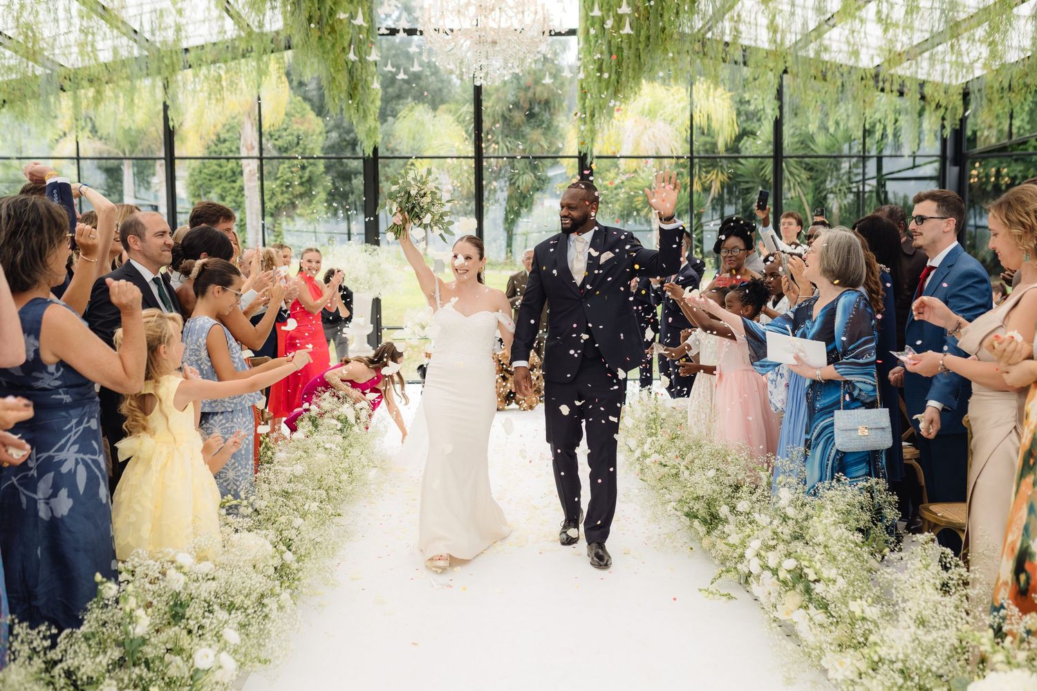 Joyful newlyweds walk down flower-lined aisle as guests toss confetti in elegant glass greenhouse wedding venue.