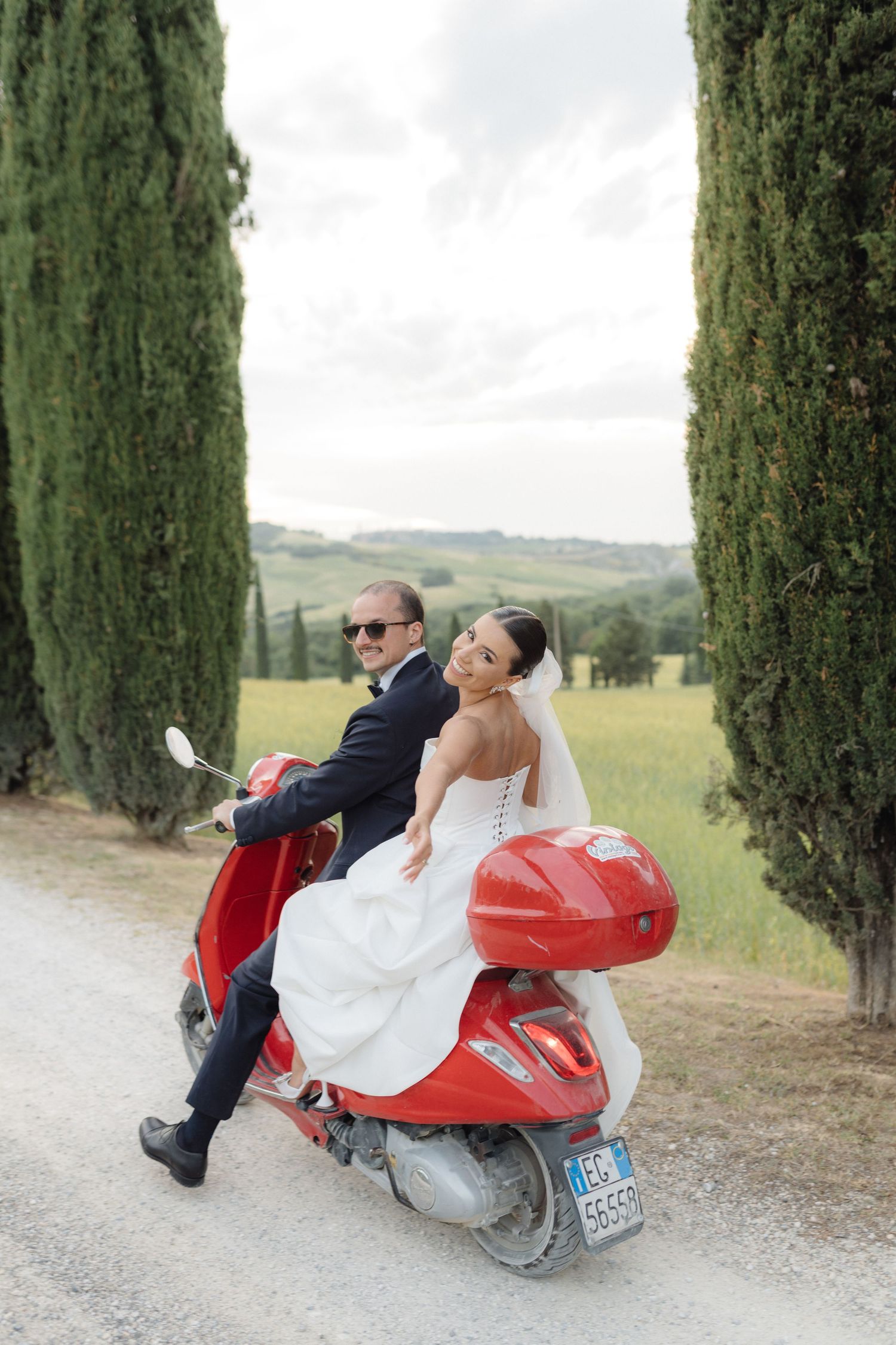 Bride and groom riding a red Vespa scooter along a cypress-lined road in Tuscany.