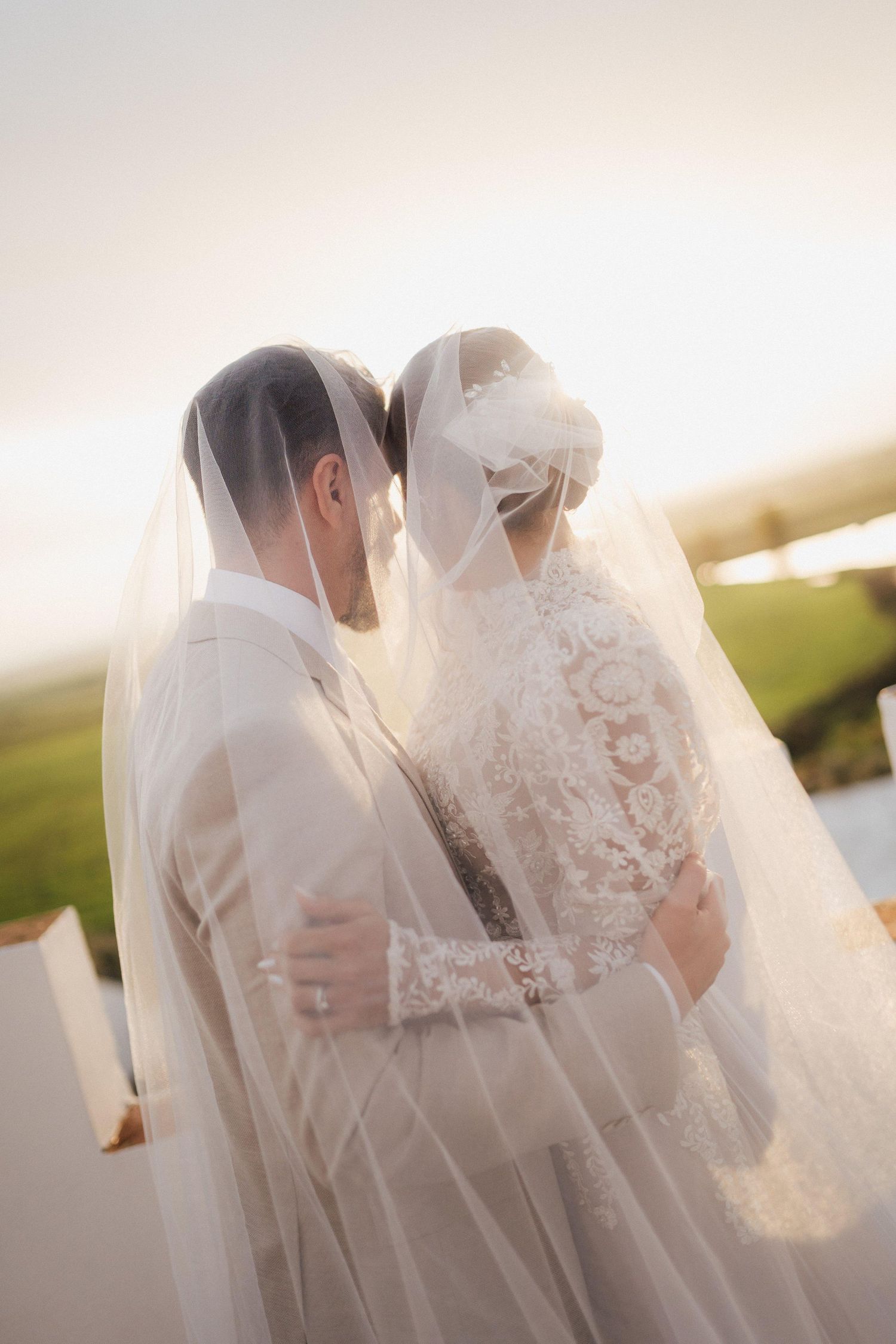 A romantic wedding couple embraces under a flowing veil during sunset.