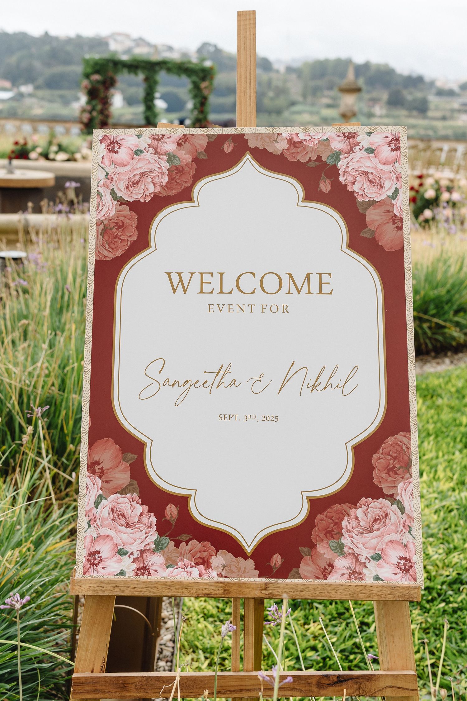 Elegant wedding welcome sign with burgundy floral design displayed on wooden easel in outdoor garden setting.