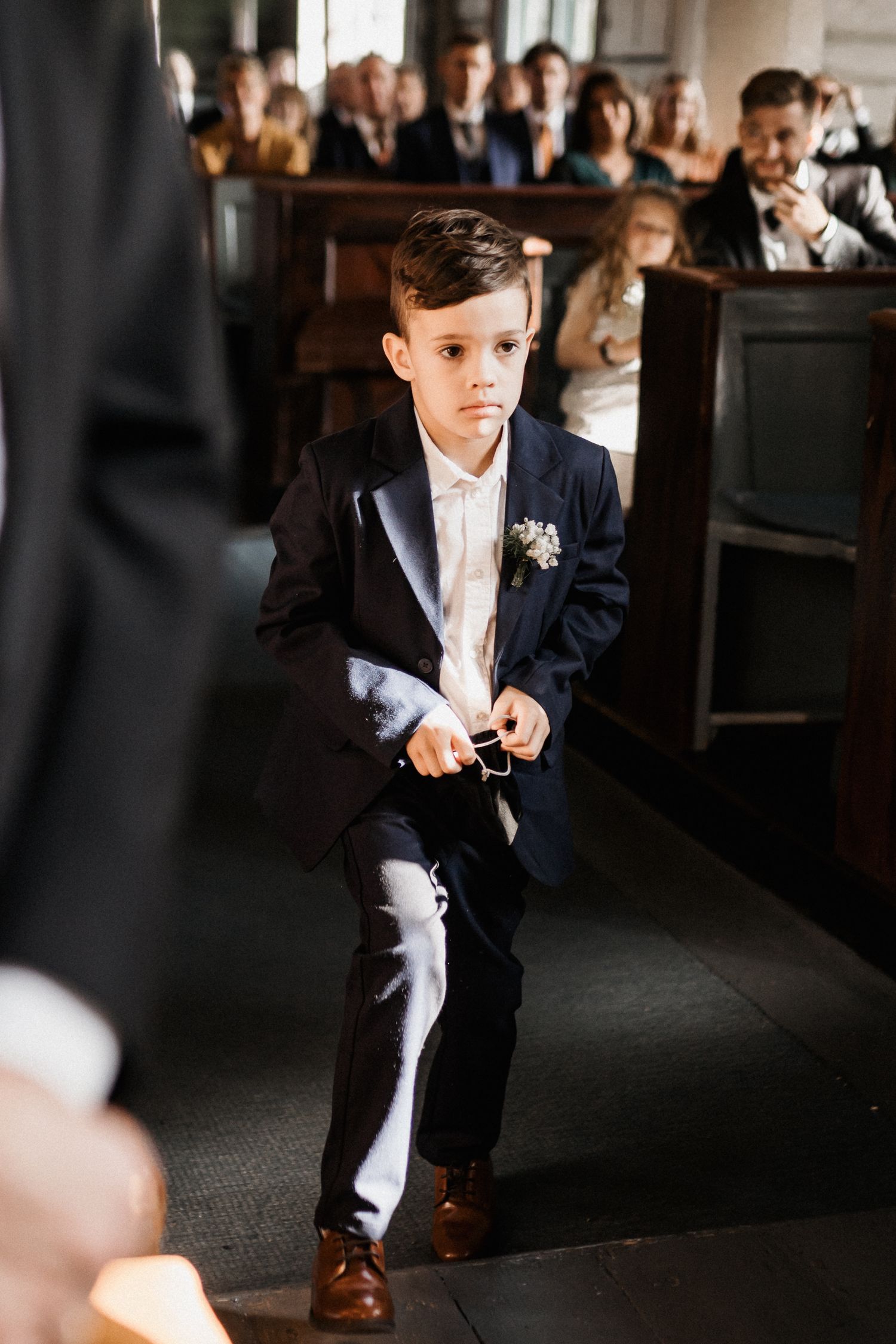 A young ring bearer in a navy suit crouches down during a wedding ceremony.