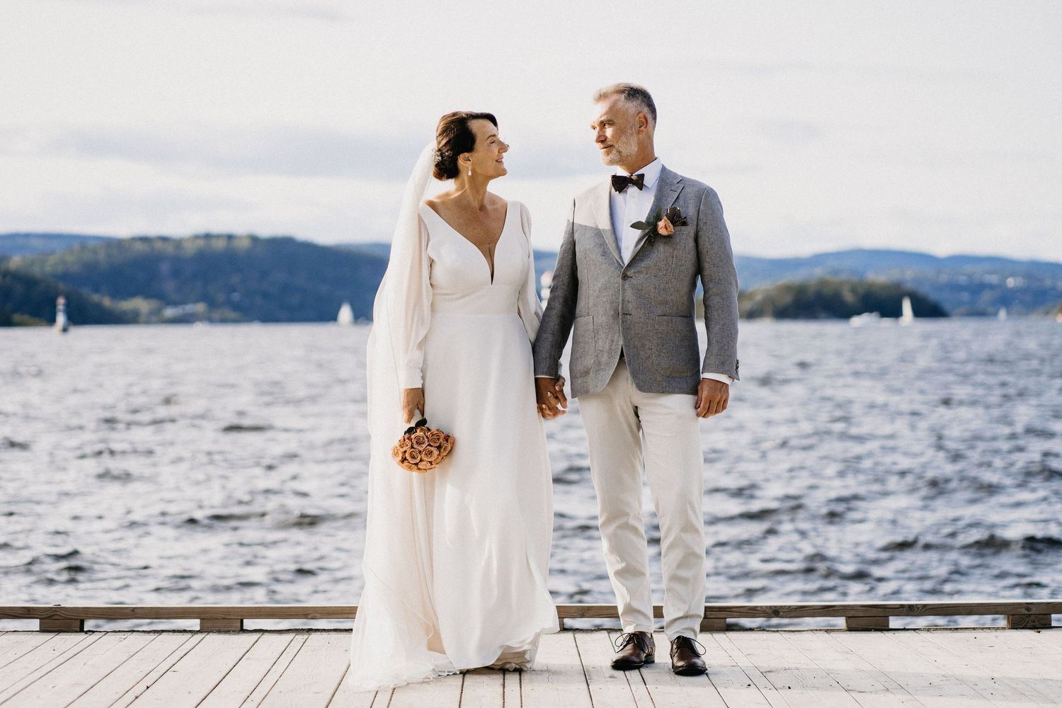 Couple in wedding attire walking along a waterfront with sailboats in the background.