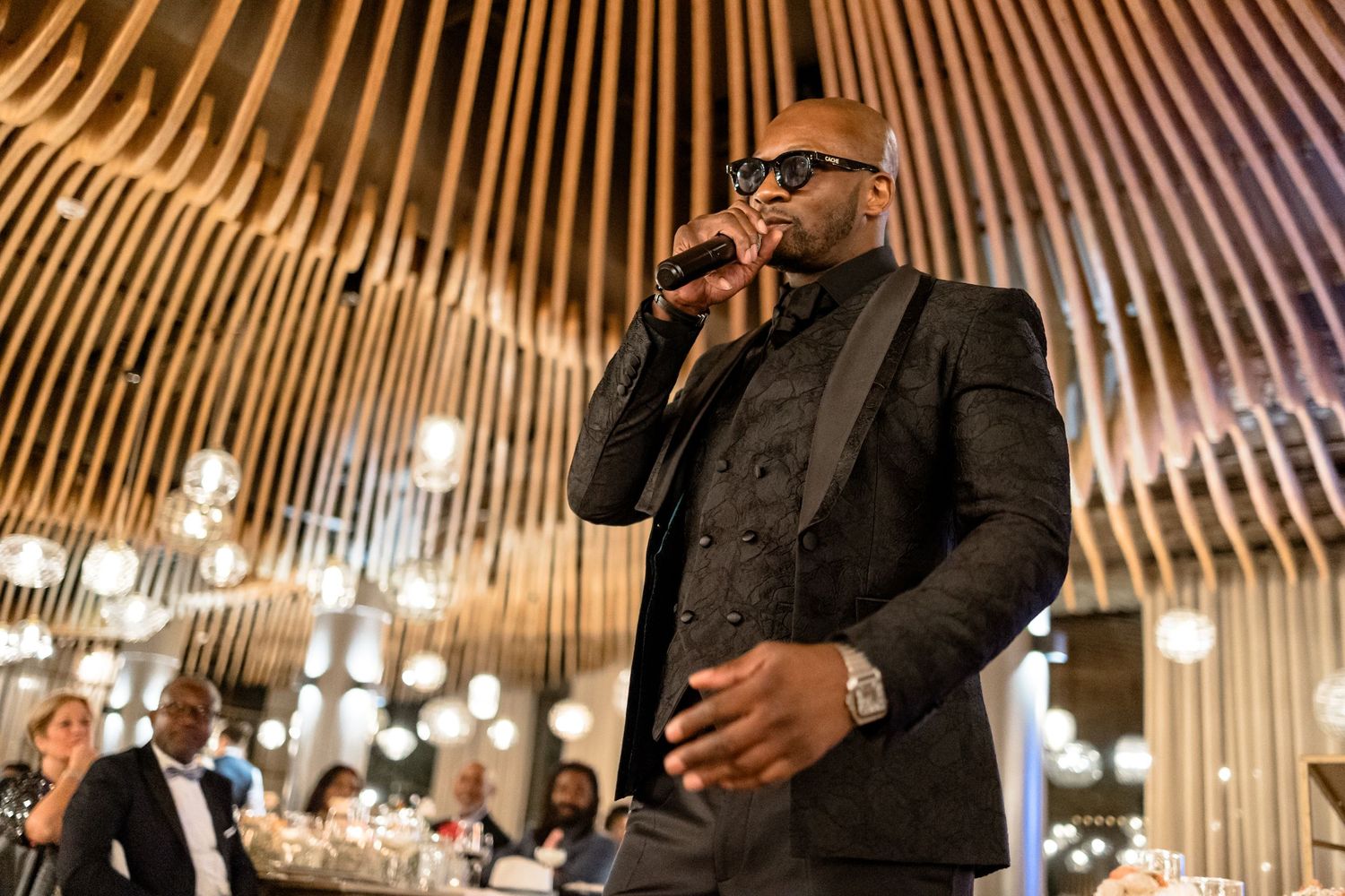 Speaker performing at wedding reception under dramatic wooden beam ceiling with pendant lights.