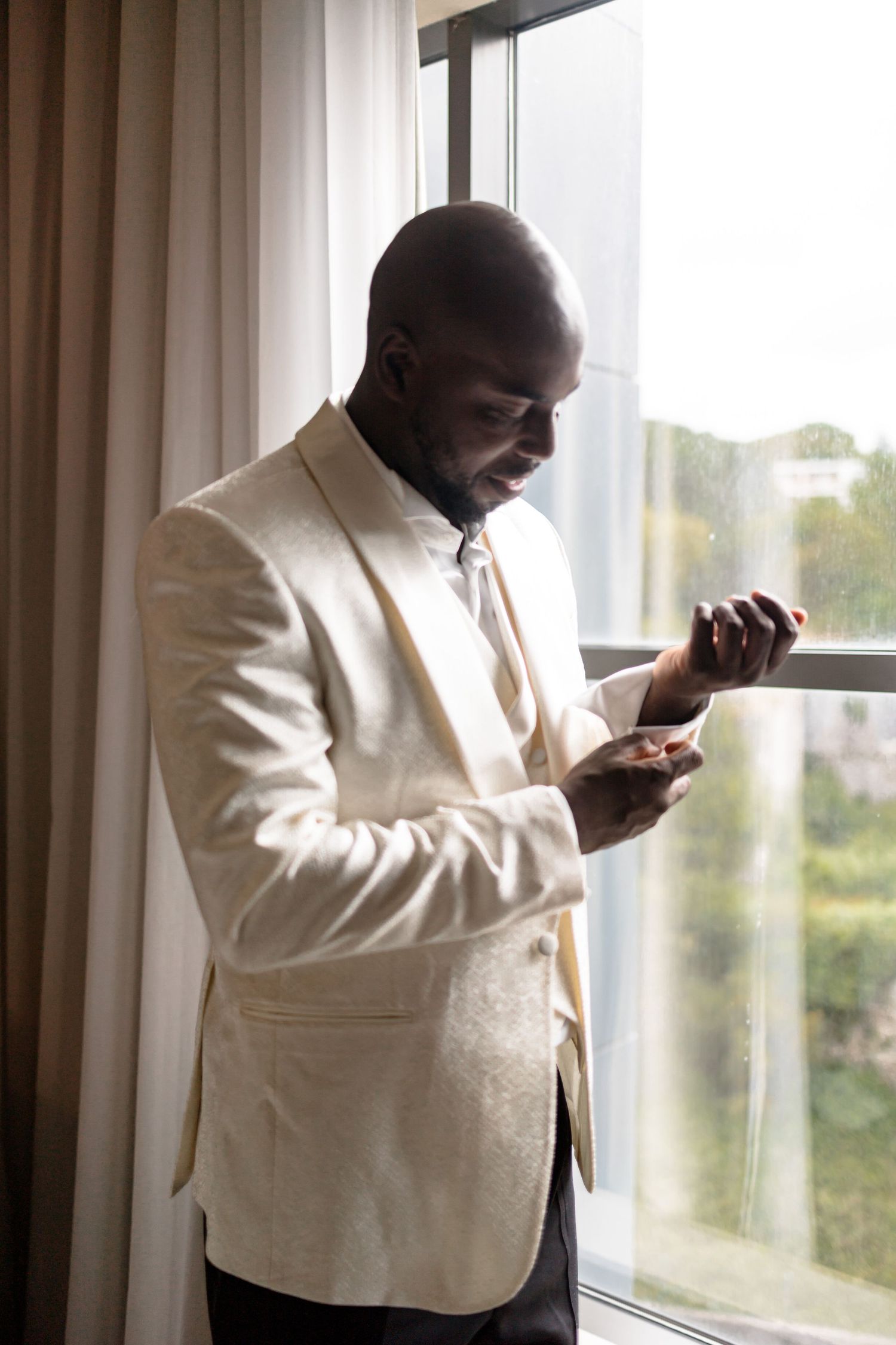 A groom in a cream tuxedo jacket checks his watch while standing by a window.