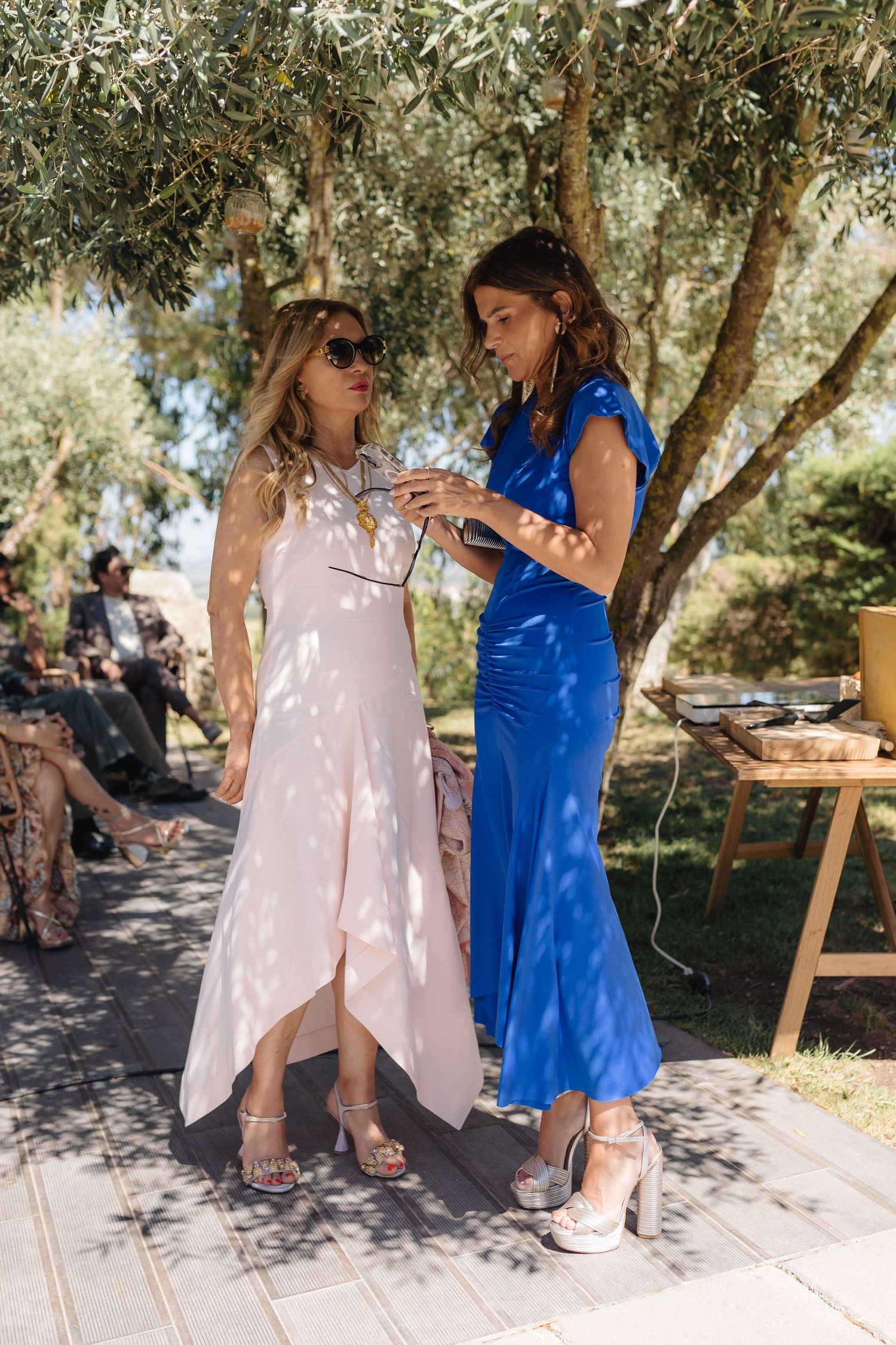 Two people in blue and pink dresses chatting under olive trees at an outdoor gathering.