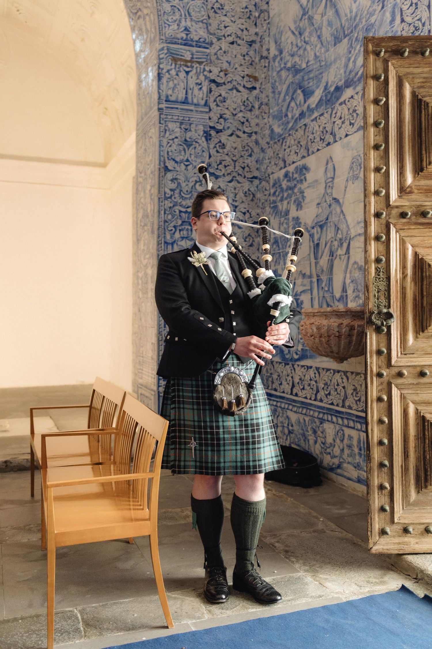 Bagpiper in traditional Scottish attire performs in a room with blue tiled walls.