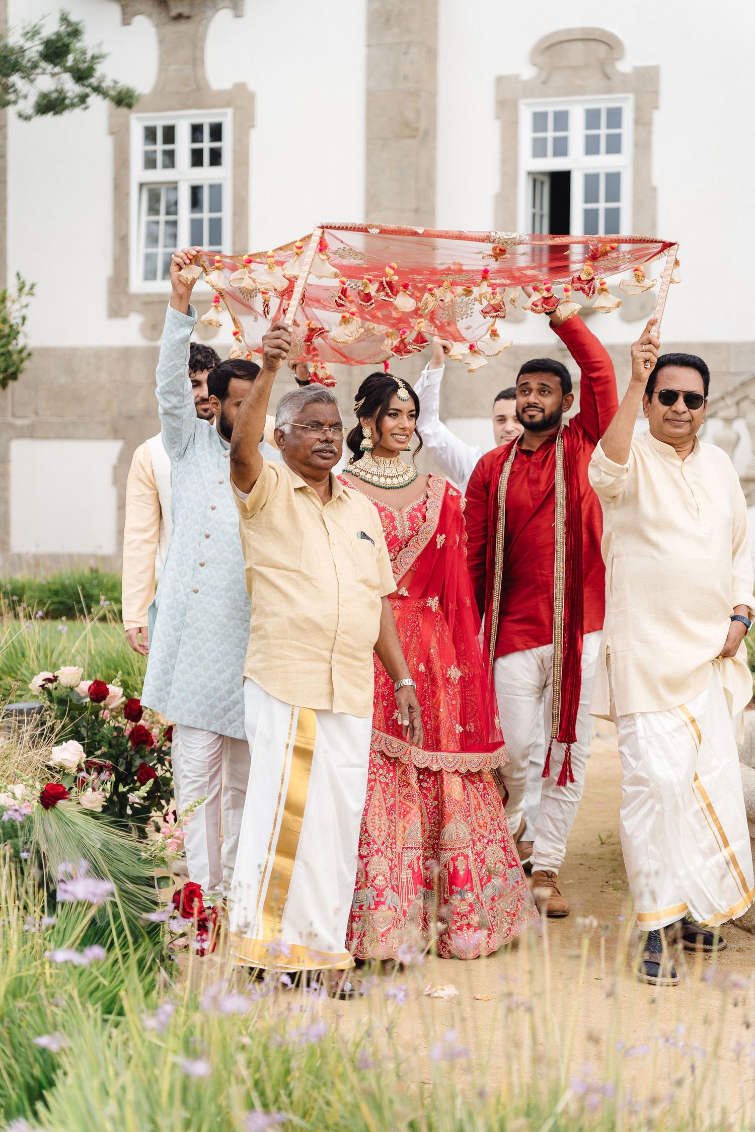 Wedding party members in traditional Indian attire walk together through a garden setting.