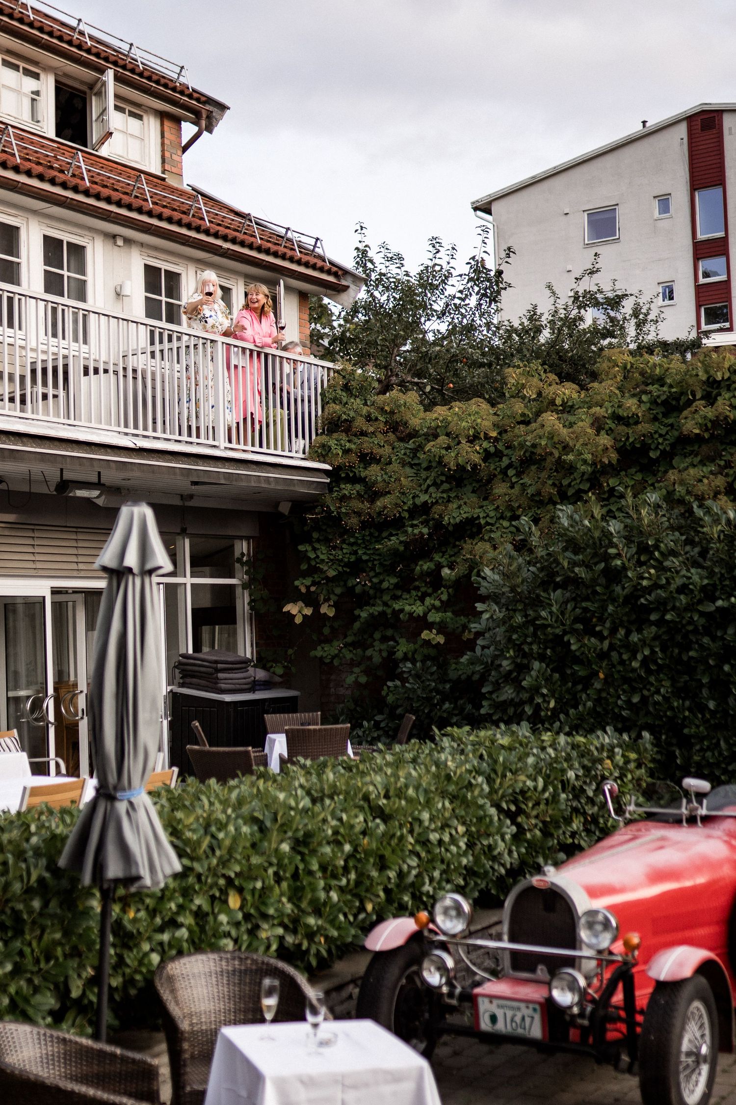 A vintage red car parked outside residential buildings with balconies and shrubs.