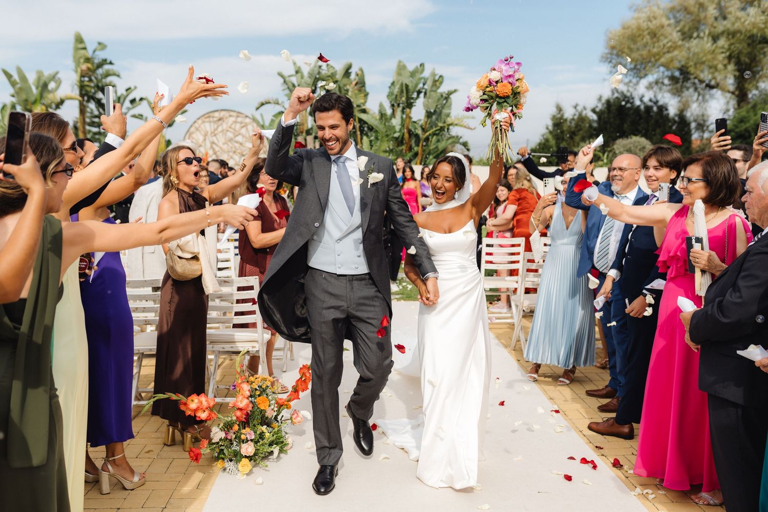 Joyful newlyweds walk down the aisle as guests celebrate with flower petals at an outdoor wedding ceremony.