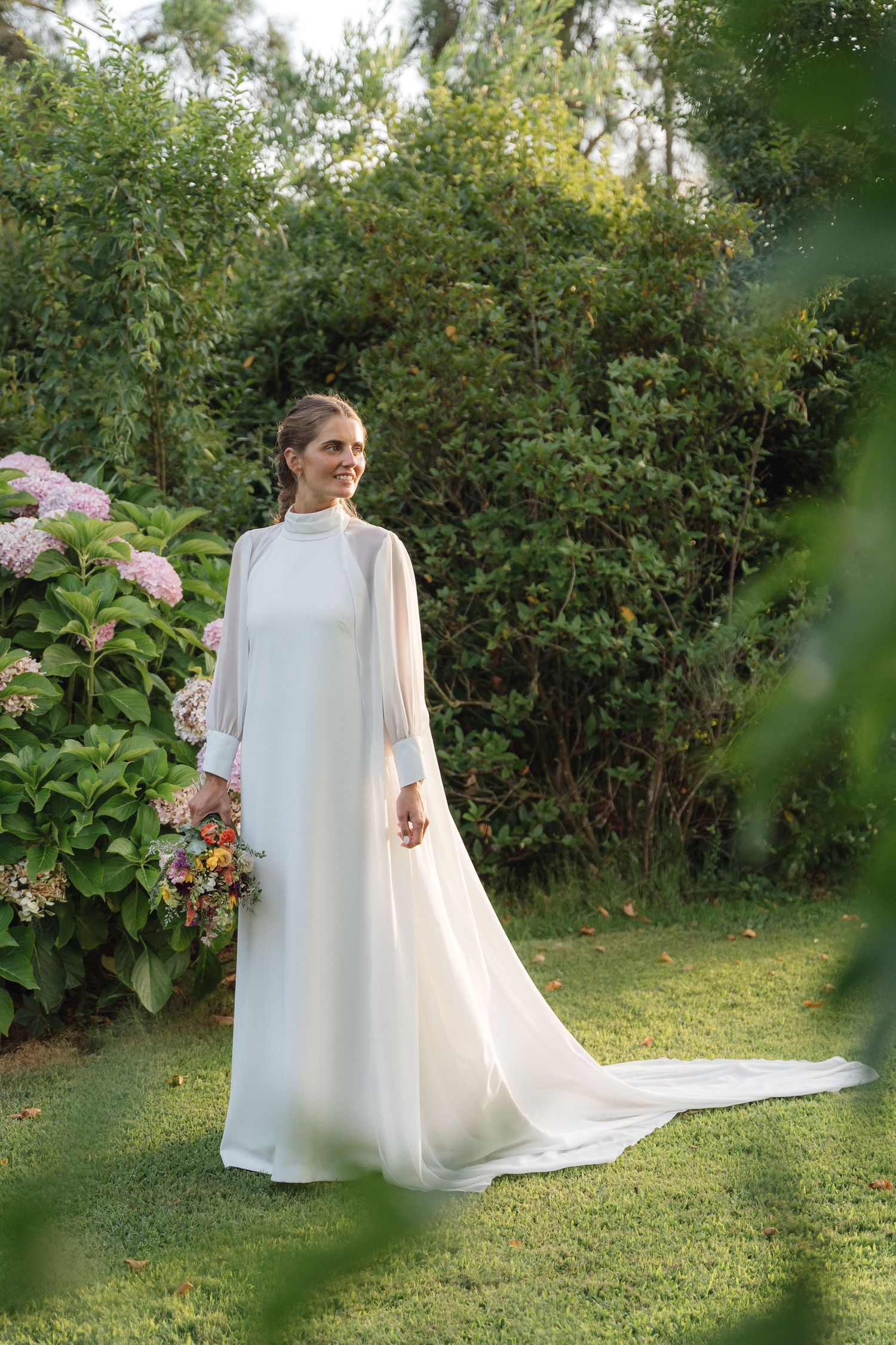 Wedding dress and cape flow elegantly in a garden setting with lush greenery.