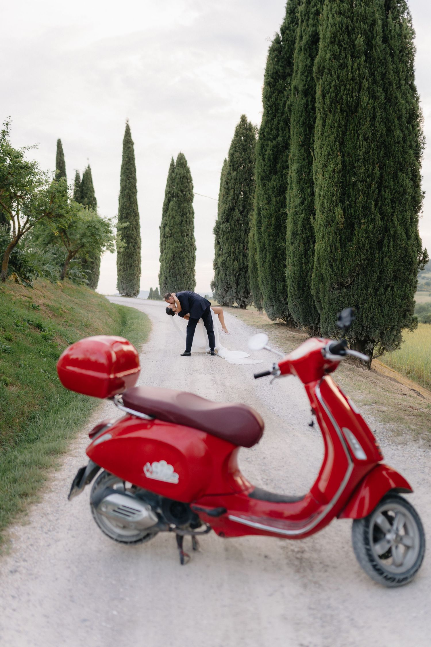 Red Vespa scooter in foreground as groom dips bride on a cypress-lined Tuscan gravel road.
