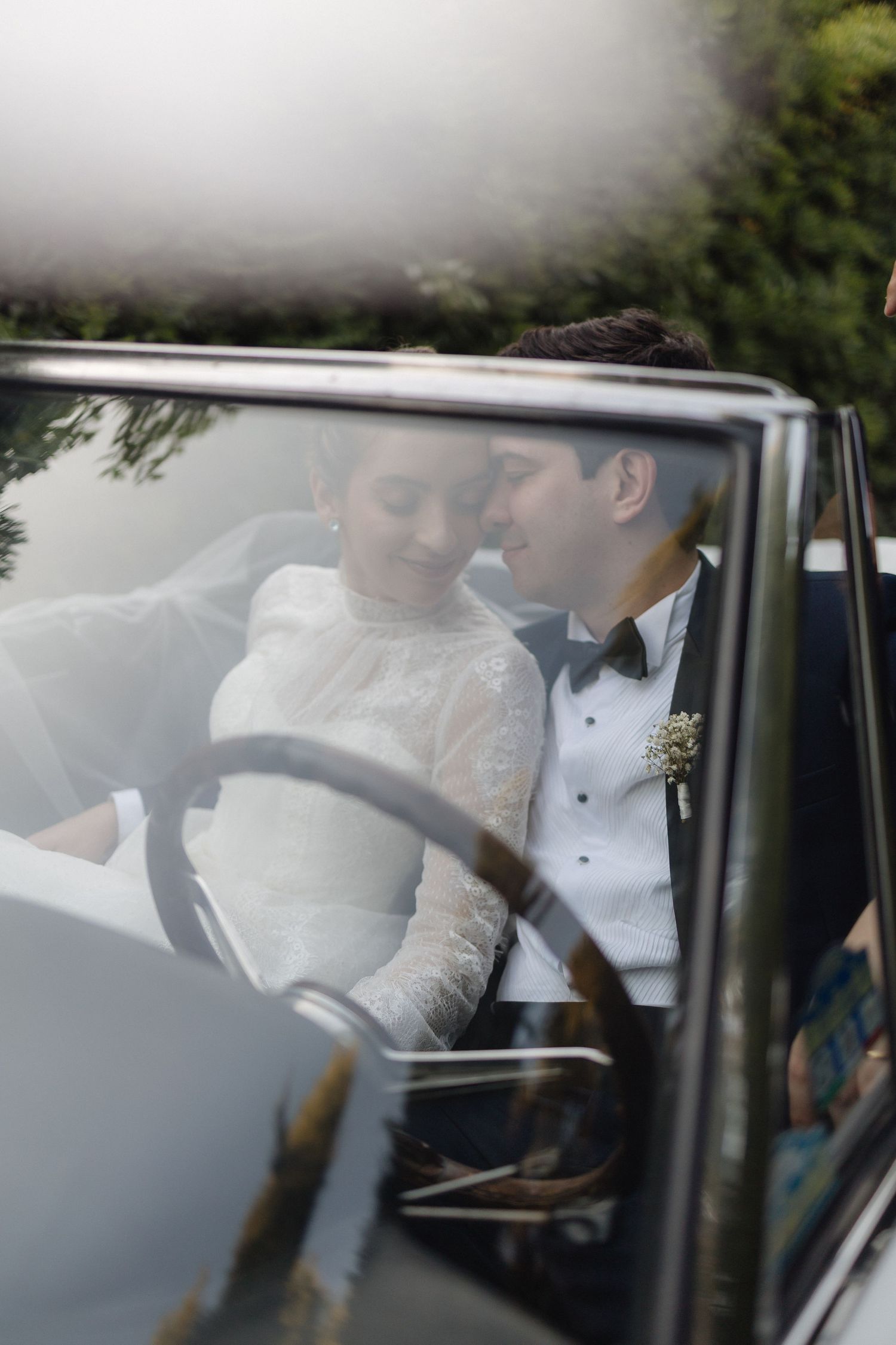 Bride and groom share an intimate moment inside a vintage car, viewed through the windshield on their wedding day.