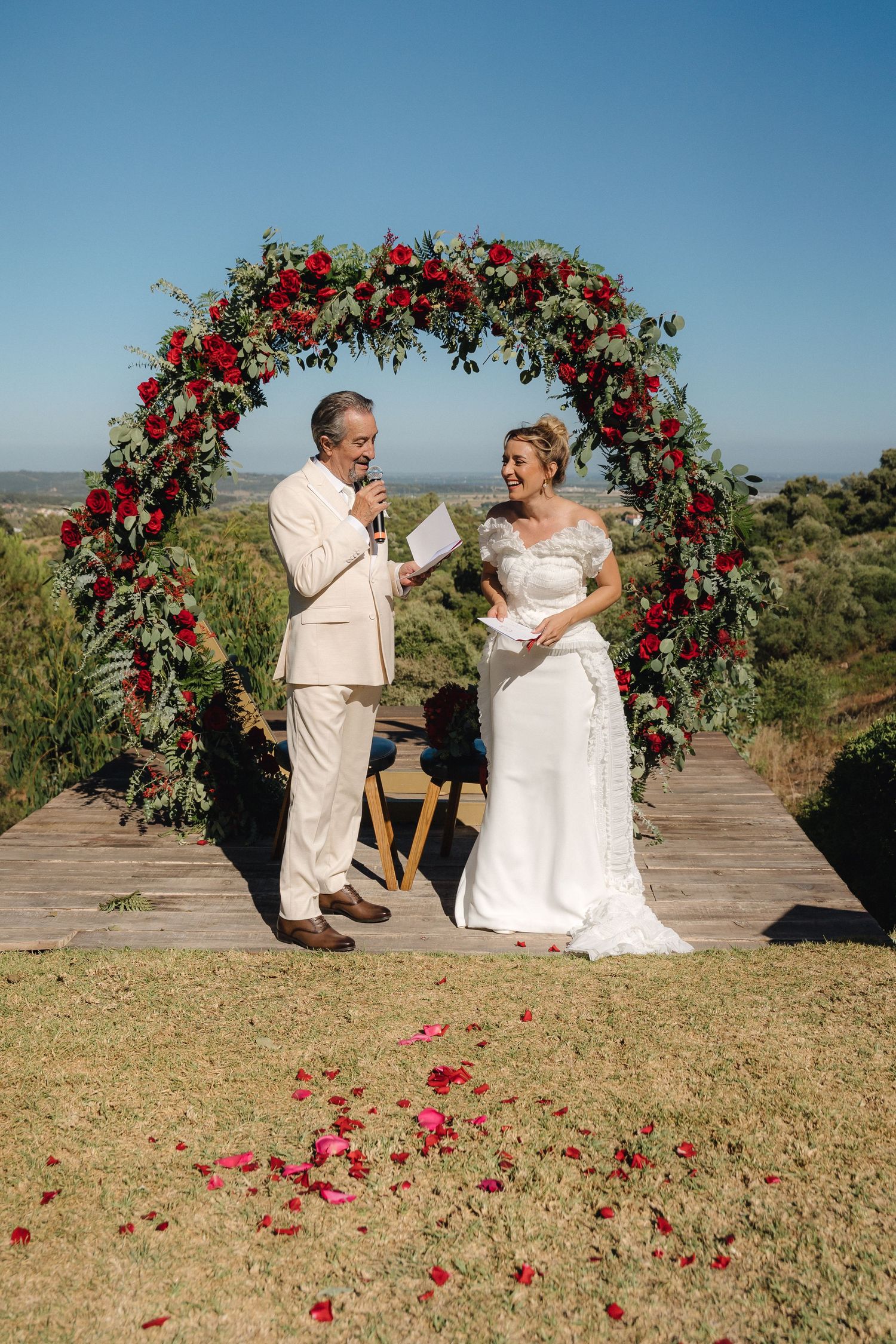 A beautiful outdoor wedding ceremony under a red floral arch with a scenic mountain backdrop.