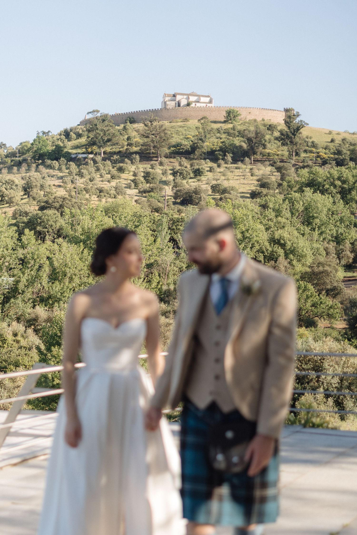 Wedding couple poses on terrace with Mediterranean hillside and fortress in background.