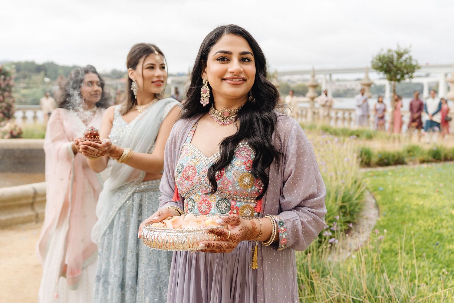 Wedding guests in pastel traditional attire carrying ceremonial items outdoors.