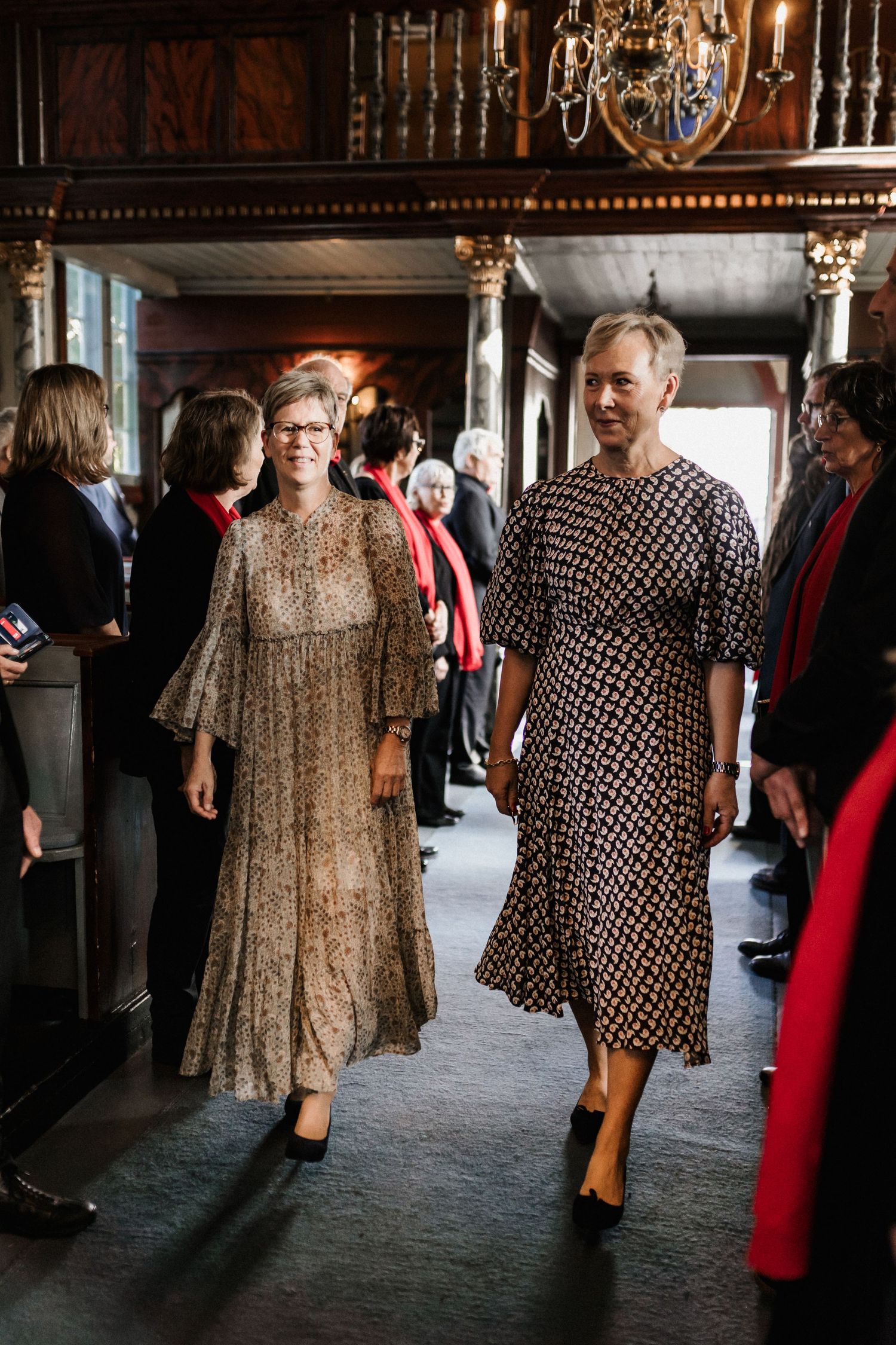 Two guests in patterned dresses walk together through an elegant hallway with ornate wooden details.