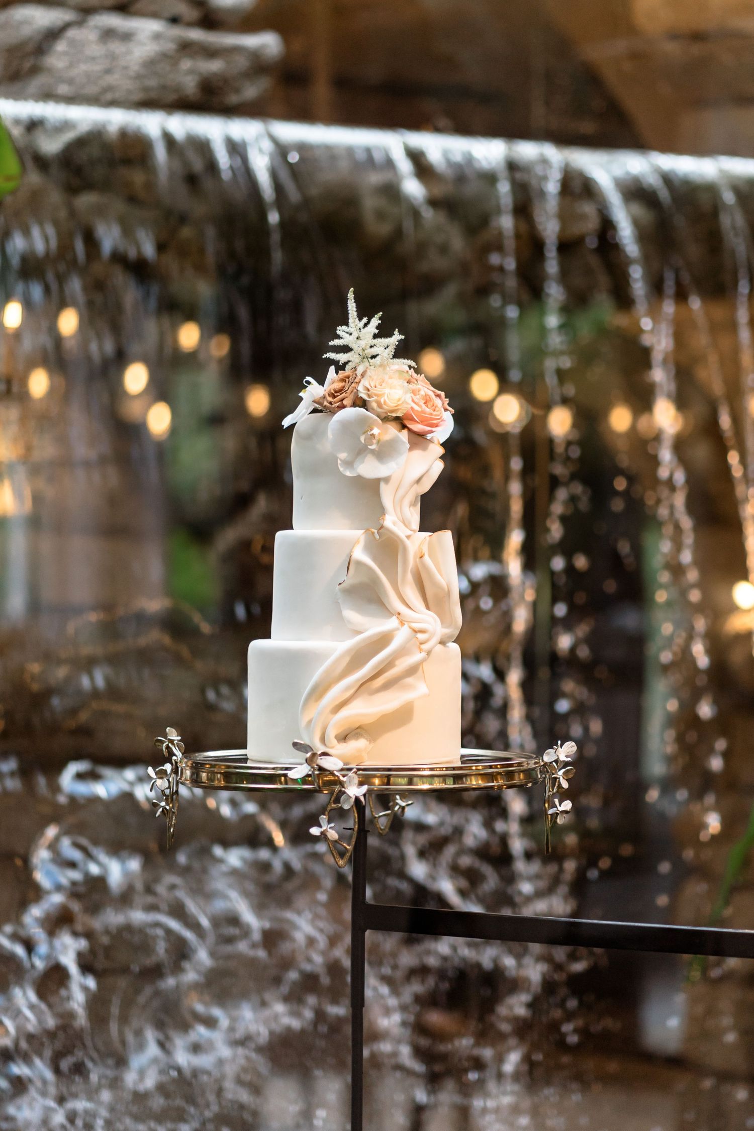 A modern white wedding cake with blush draping and floral accents displayed against a waterfall backdrop.