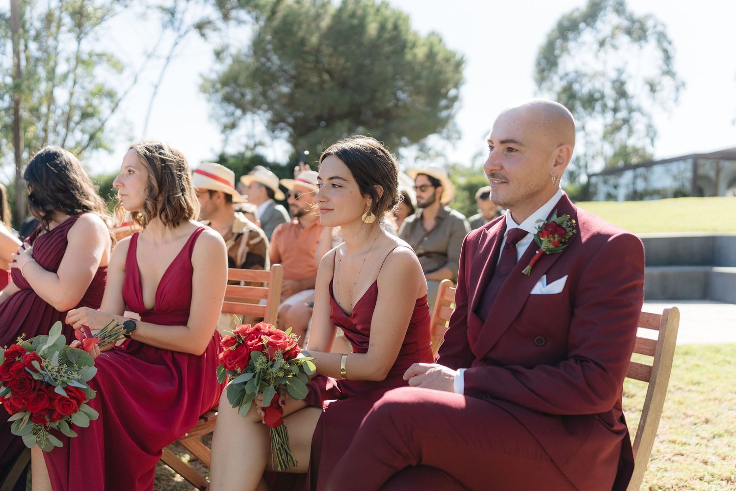 Wedding party in burgundy attire seated during outdoor ceremony.