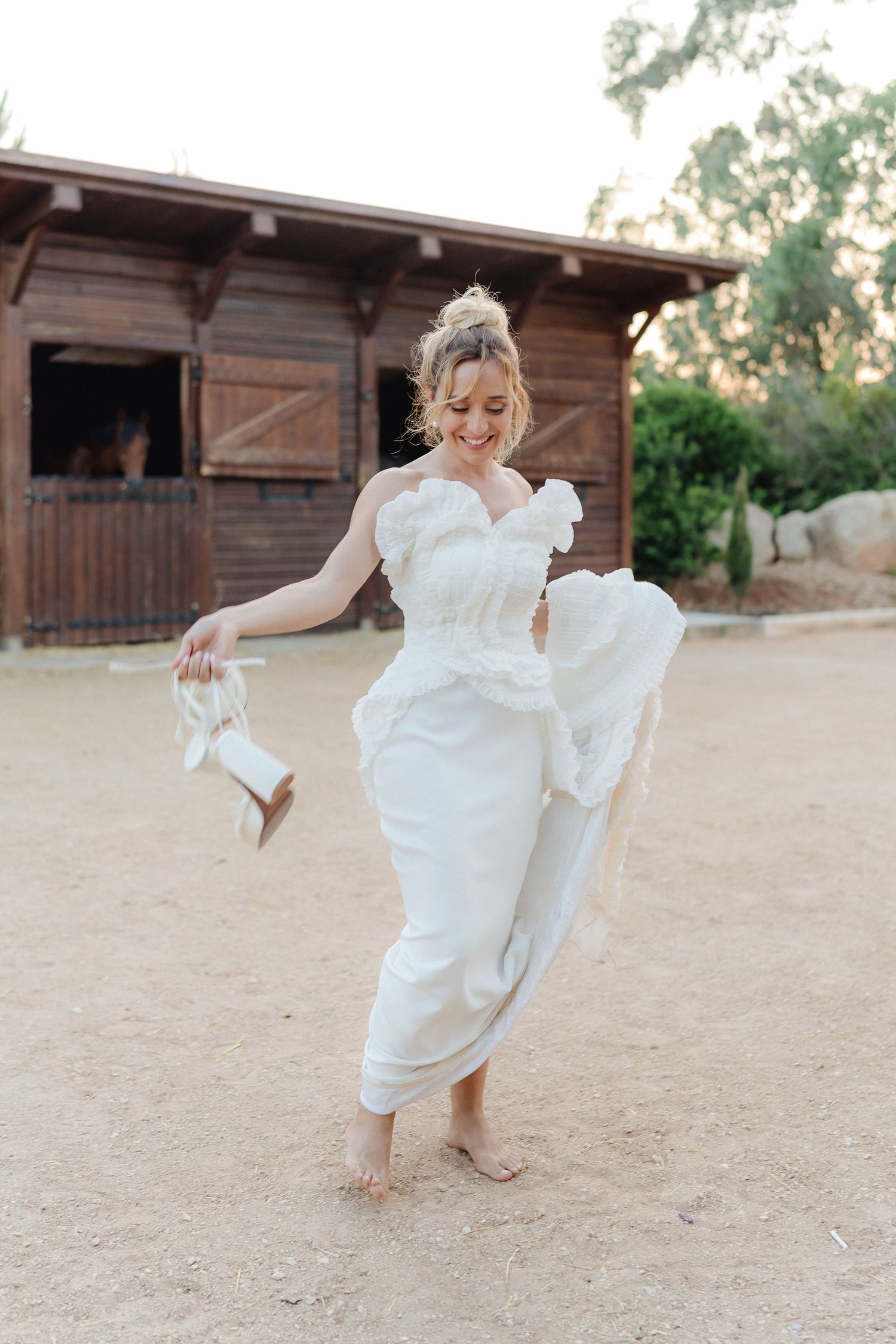 A person in a white dress dances joyfully on sandy ground near wooden stables.
