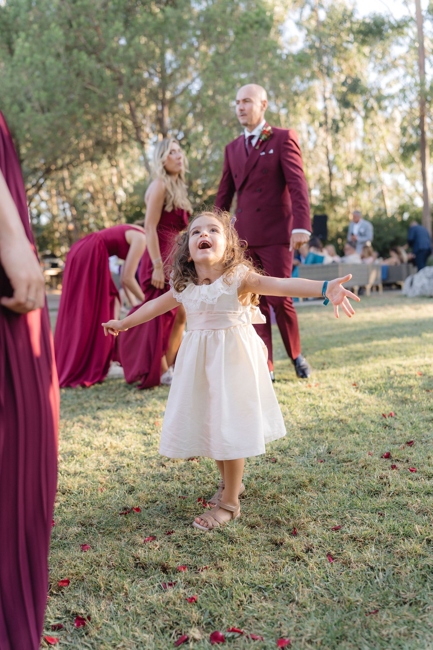 Young flower girl in white dress dances joyfully at outdoor wedding with burgundy-dressed attendants.