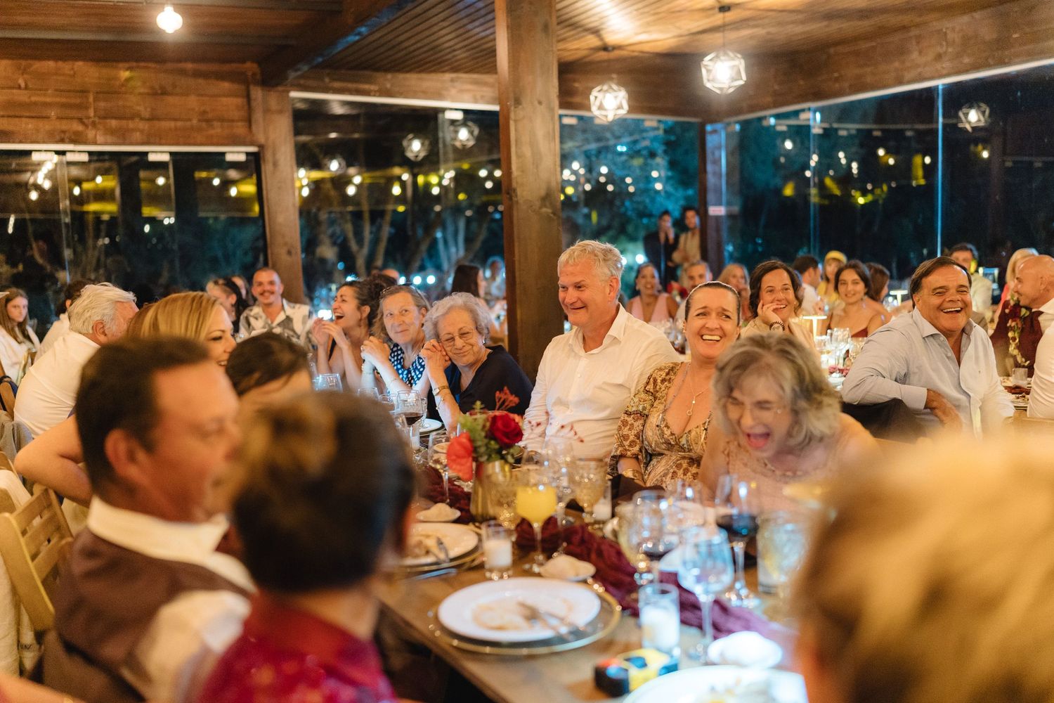 Wedding reception guests seated at long tables in a rustic wooden venue with string lights.