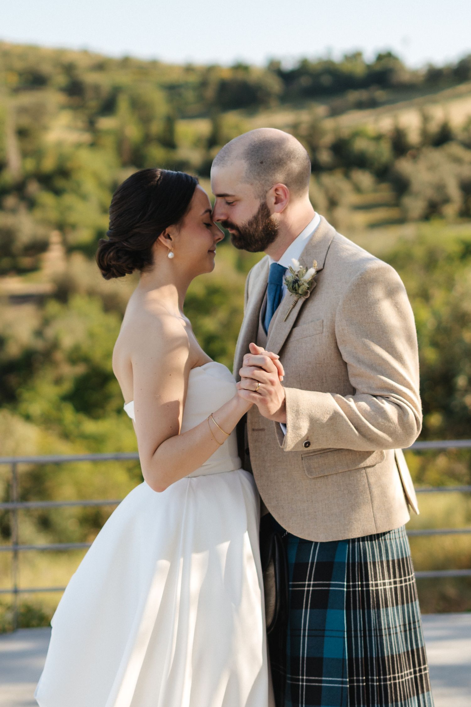 Romantic wedding couple embraces on outdoor terrace with scenic hillside view.