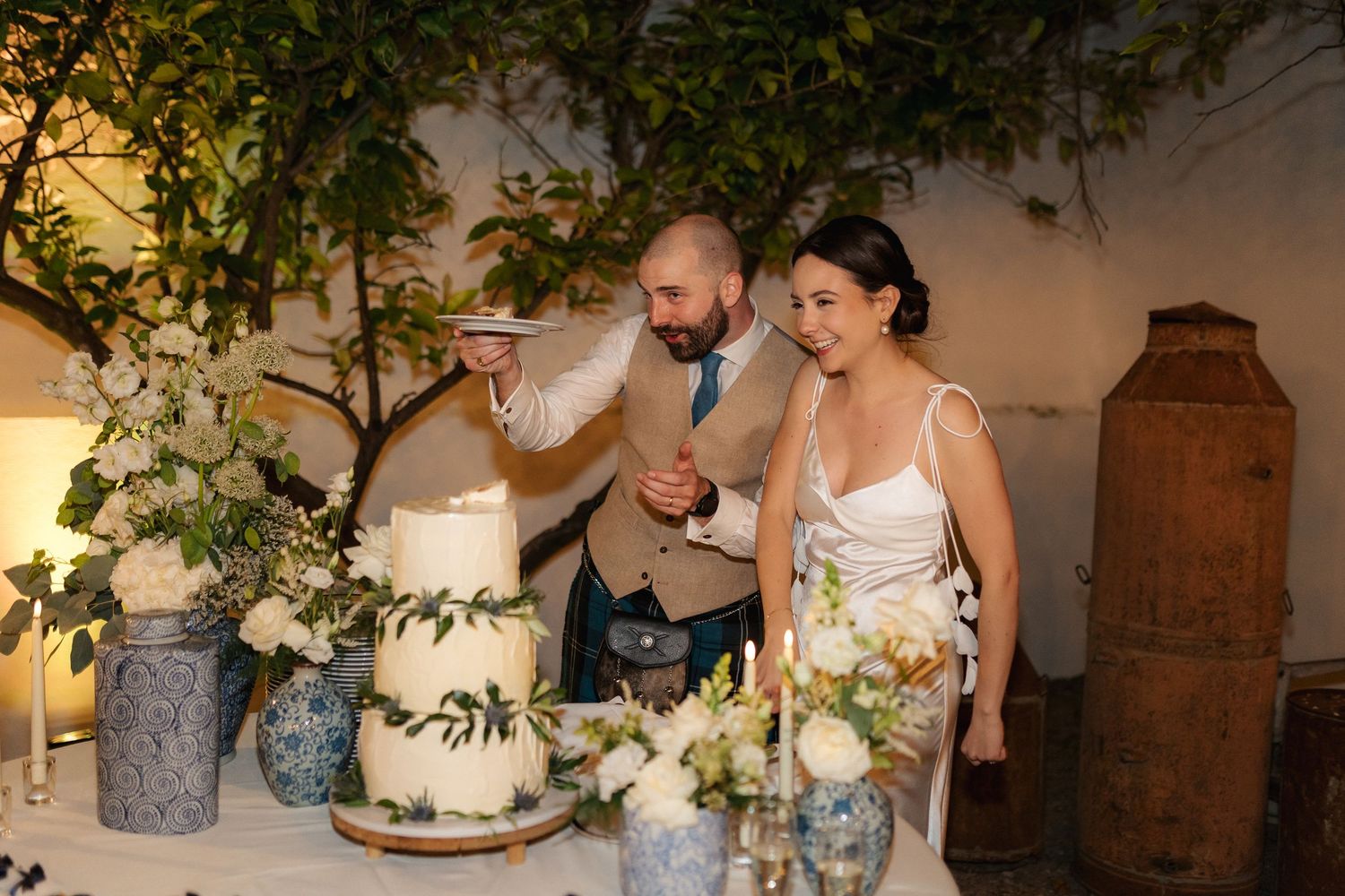 Couple cutting their elegant white wedding cake decorated with greenery.