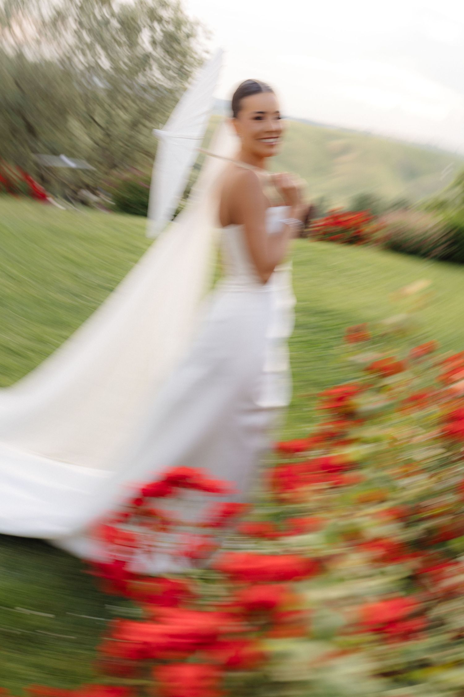 Smiling bride in white gown and veil walking through red poppy flower garden on wedding day.
