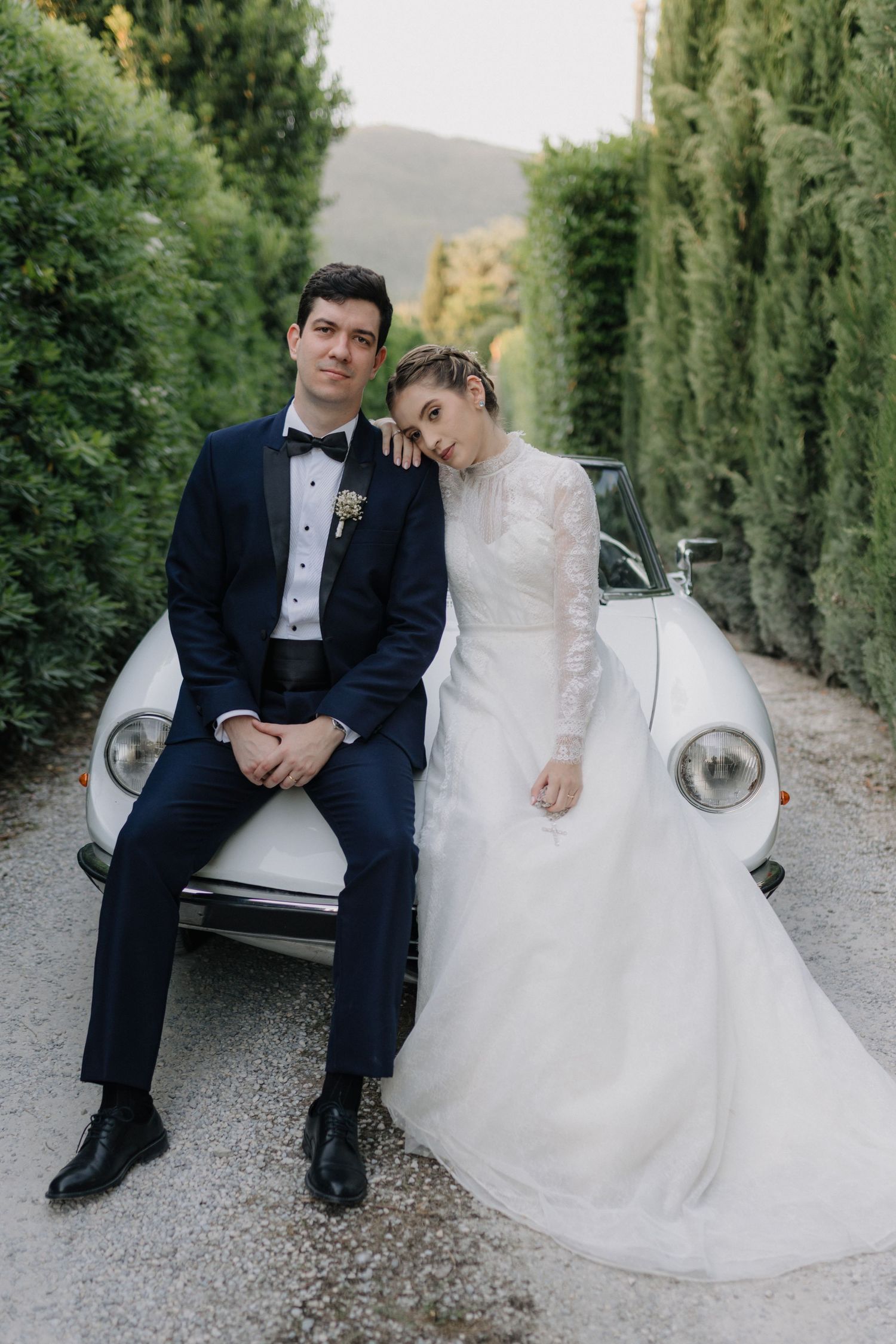 Elegant bride and groom posing against a vintage white car on a tree-lined gravel path.