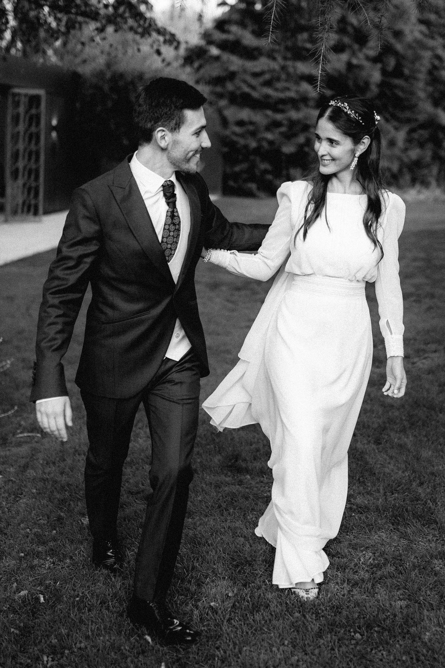 Black and white photo of playful couple running through grass in wedding attire.